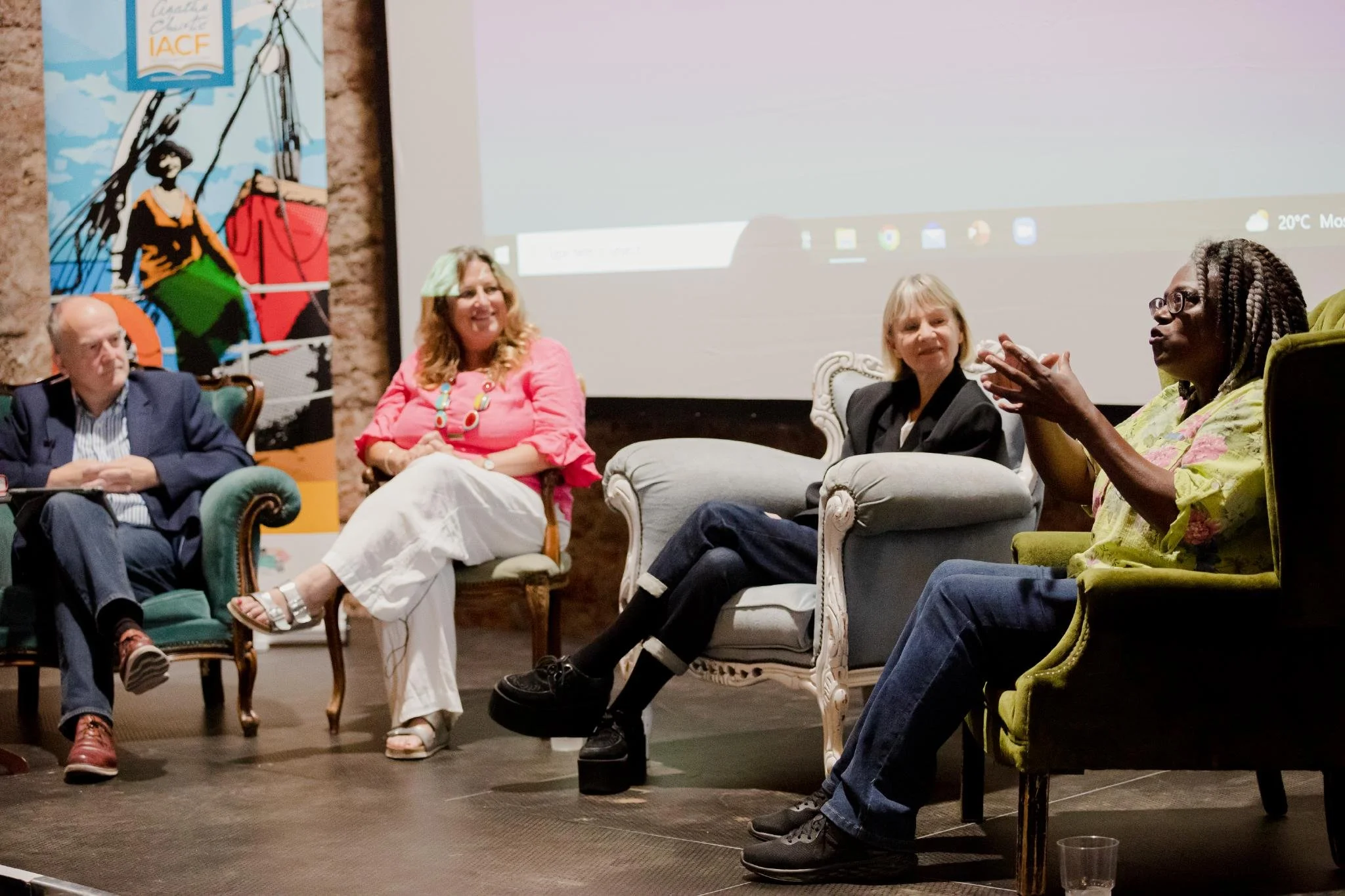 A panel discussion with five people sitting on vintage chairs and sofas on a stage, with a large screen behind them displaying a computer desktop, during a conference or workshop. A colorful mural with a woman and abstract shapes is visible on the left.