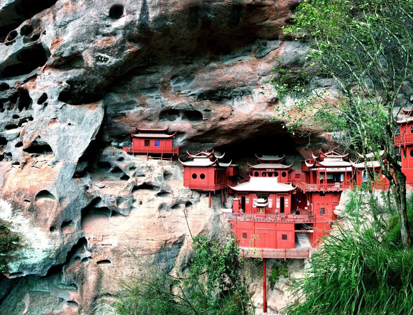 Multiple traditional Chinese temple buildings painted red, built into a rocky cliff.