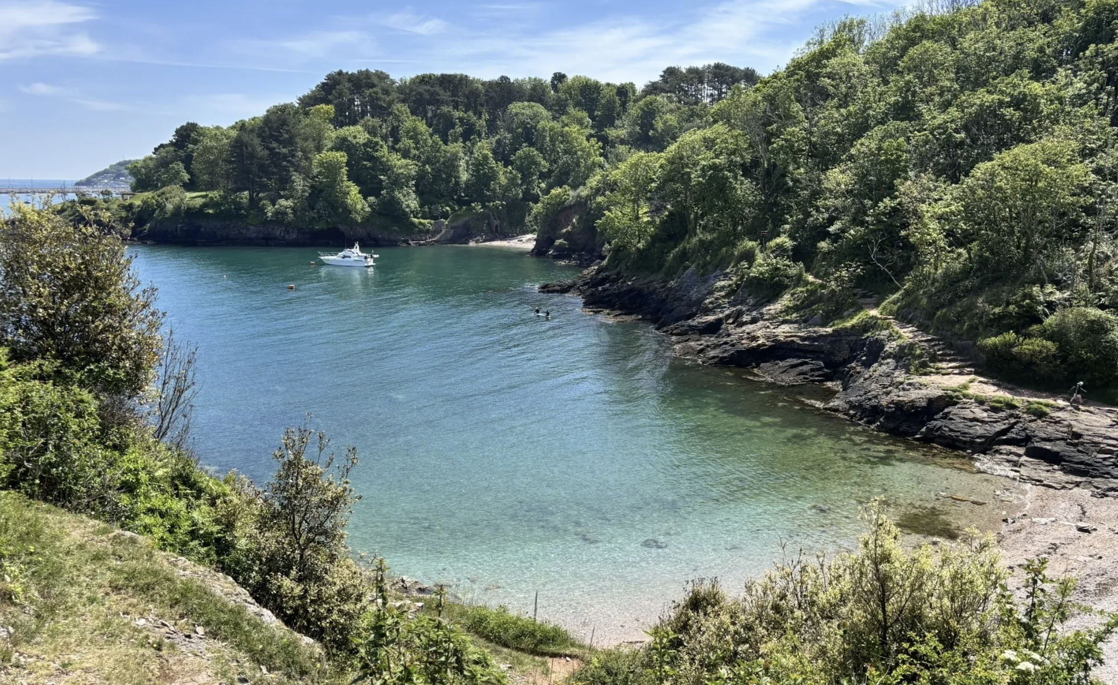 Calm water in a cove with a boat and kayakers, lush green trees covering the hills, rocky shoreline, and a blue sky with some clouds.
