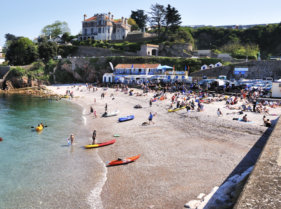 Crowded beach with people sunbathing and swimming, colorful kayaks on the shore, and a large historic house on a hill in the background.