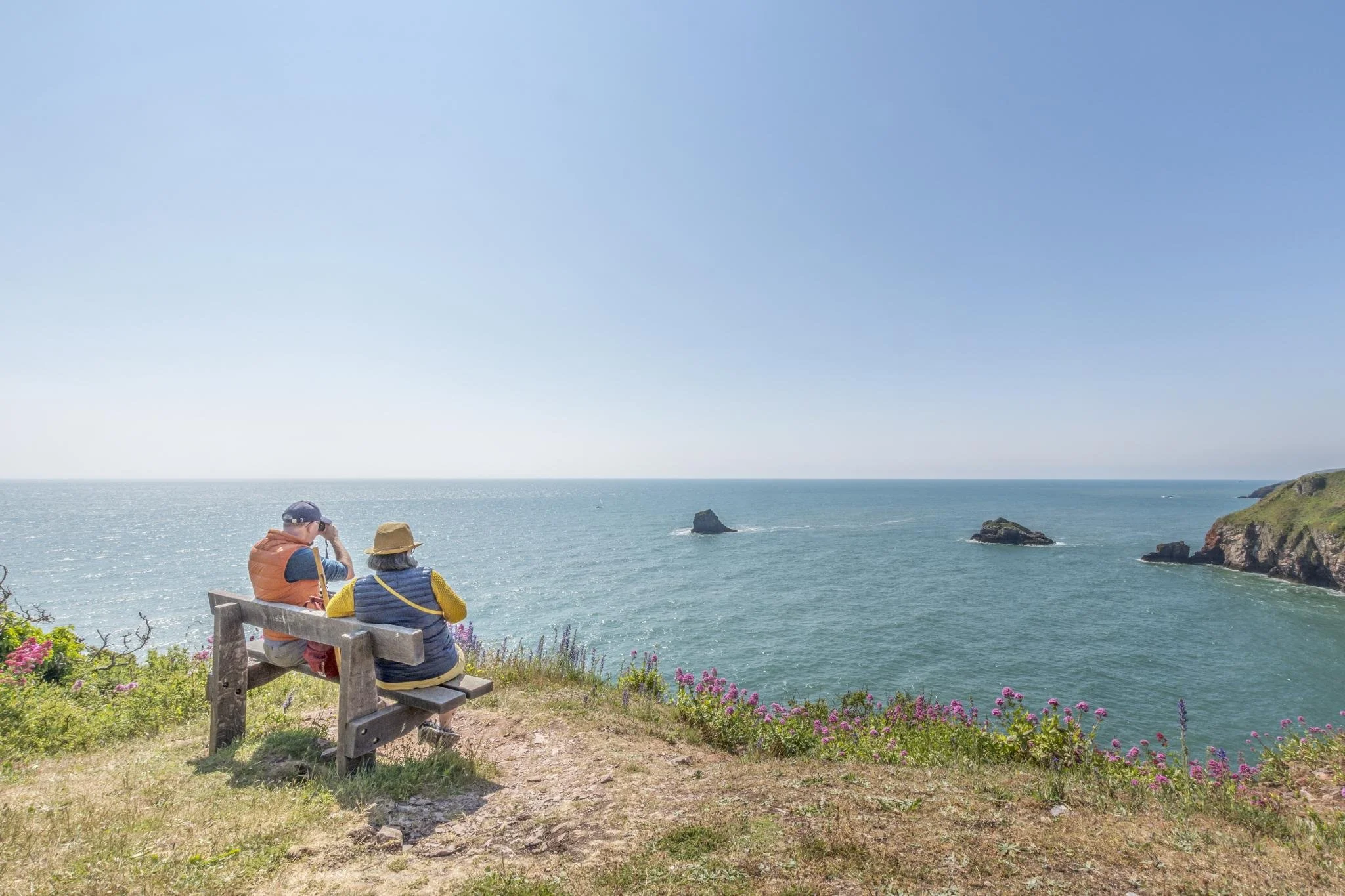 Two people sitting on a bench by the ocean, facing the water, with rocky islands and a clear blue sky in the background.