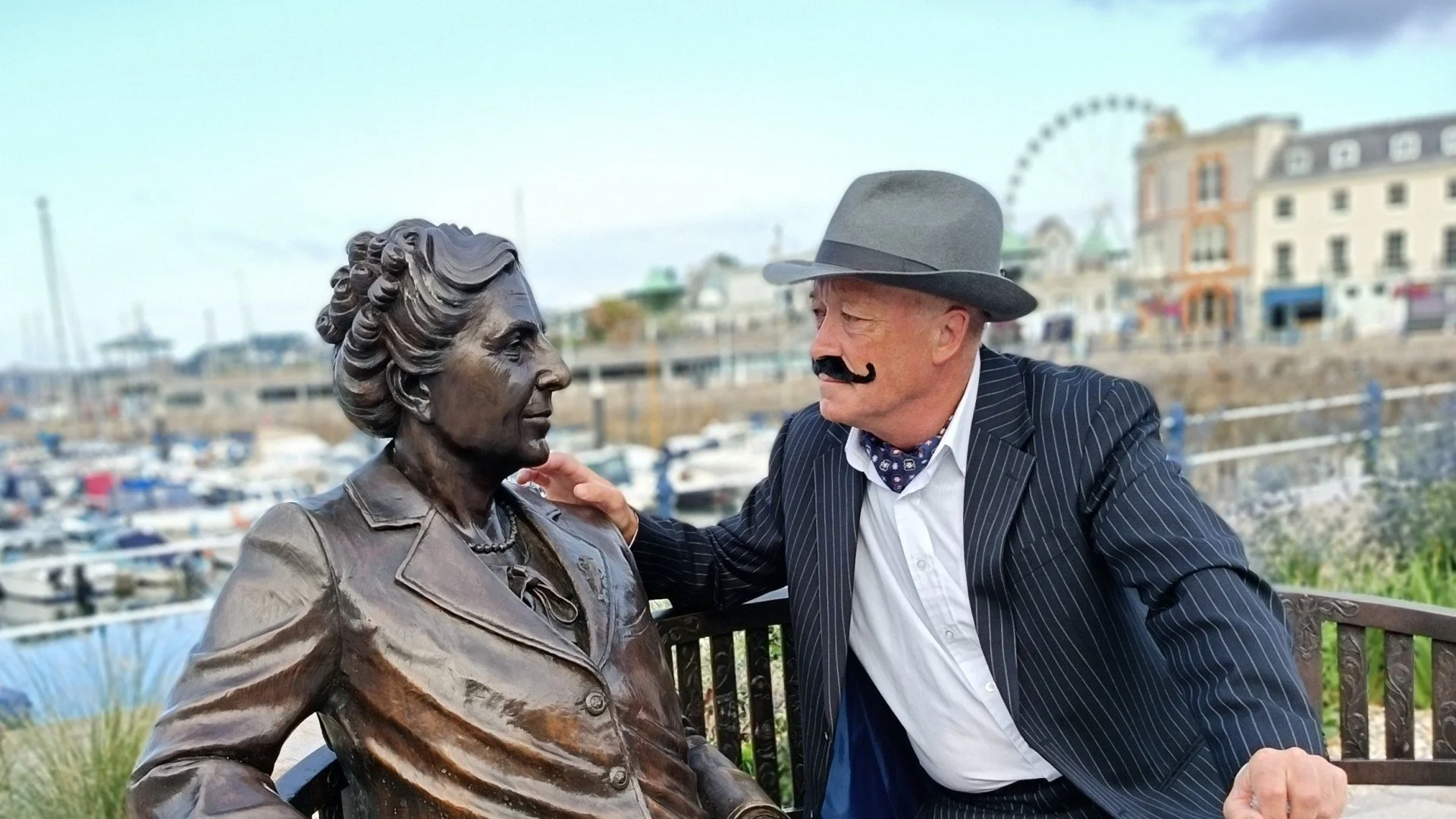 An older man with a grey fedora, striped suit, white shirt, and bandana around his neck is sitting on a bench, touching the shoulder of a bronze statue of a woman with curly hair, wearing a blazer. The scene appears to be at a waterfront with buildings and a Ferris wheel in the background.