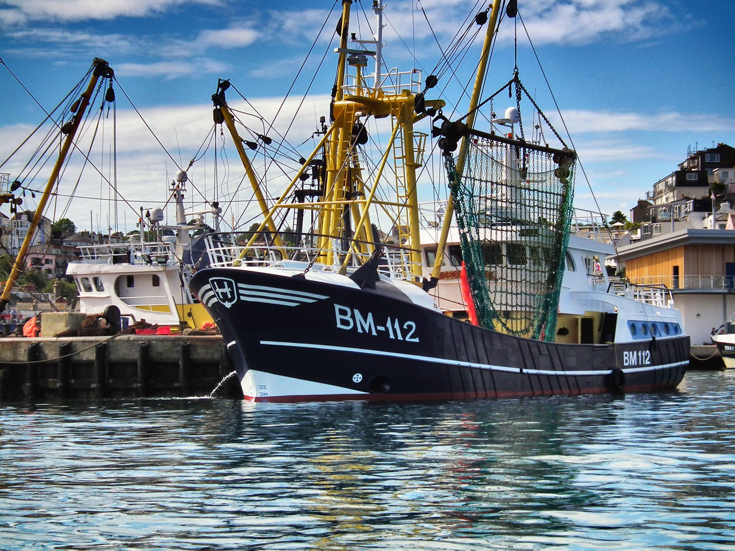 A black and white fishing boat docked at a harbor with other boats and buildings in the background.