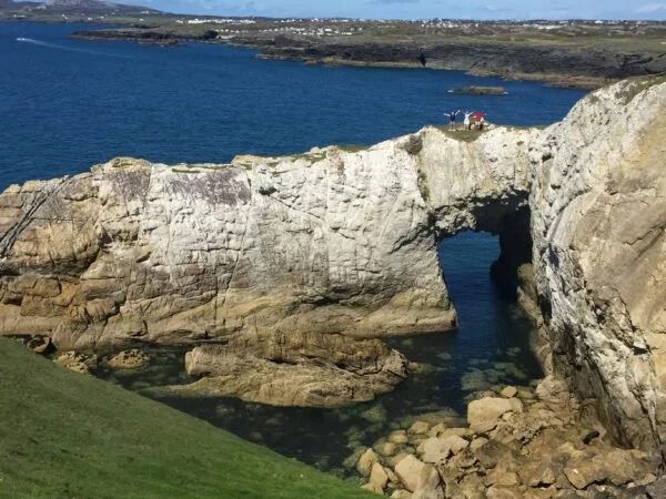Rock formation with an arch over the water, with grassy area in the foreground and coastline in the background.