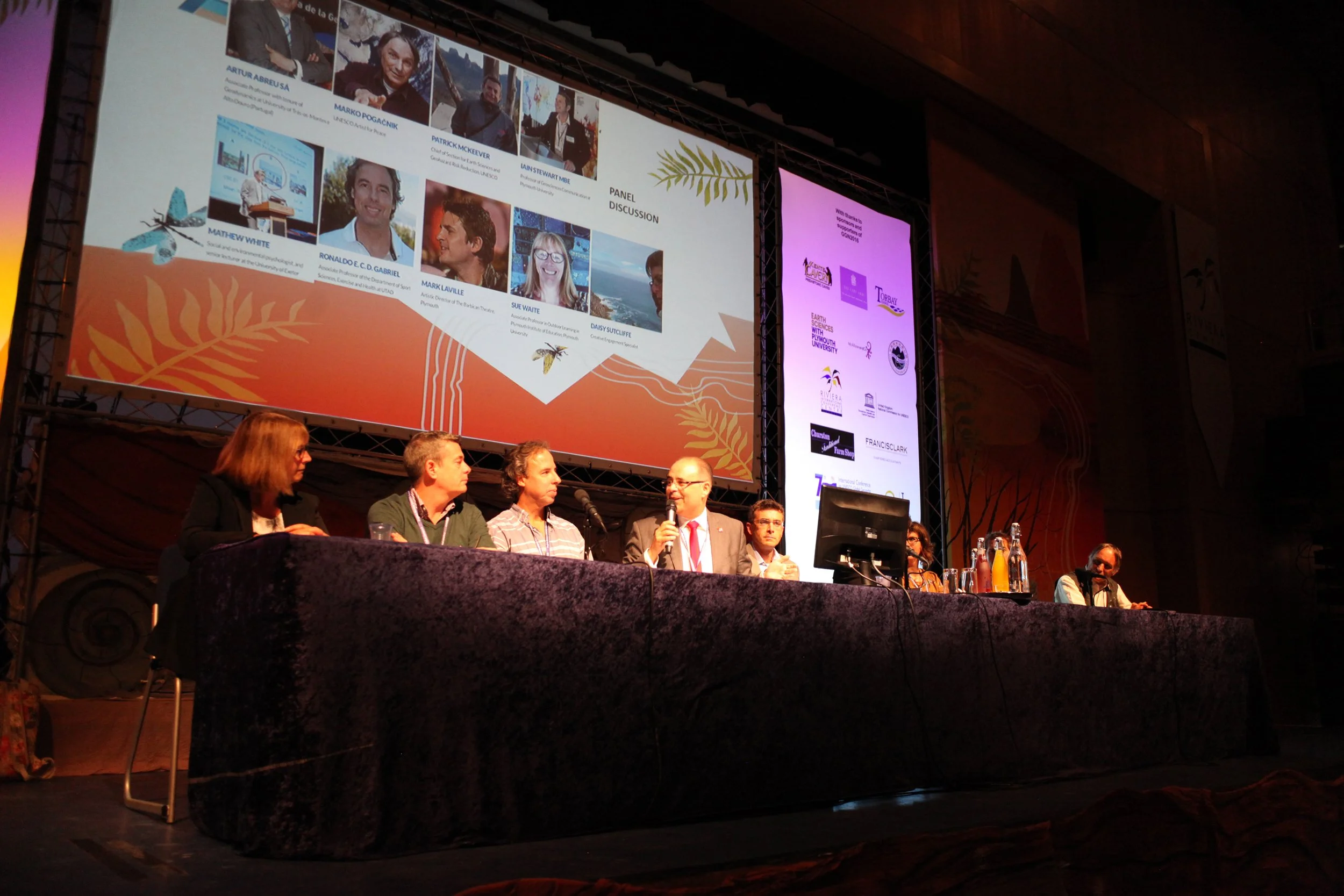 Panel discussion at a conference with seven individuals seated at a long table on stage, with a large screen behind them displaying images and text, including titles and names of participants and sponsors.