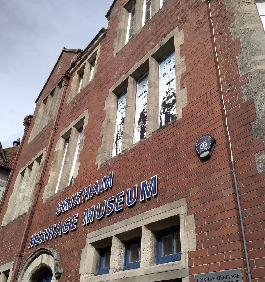 Facade of the Brixham Heritage Museum building made of red bricks with blue framed windows and white sign that reads 'BRIXHAM HERITAGE MUSEUM' on the front wall. Two posters are visible in the windows on the upper floor.