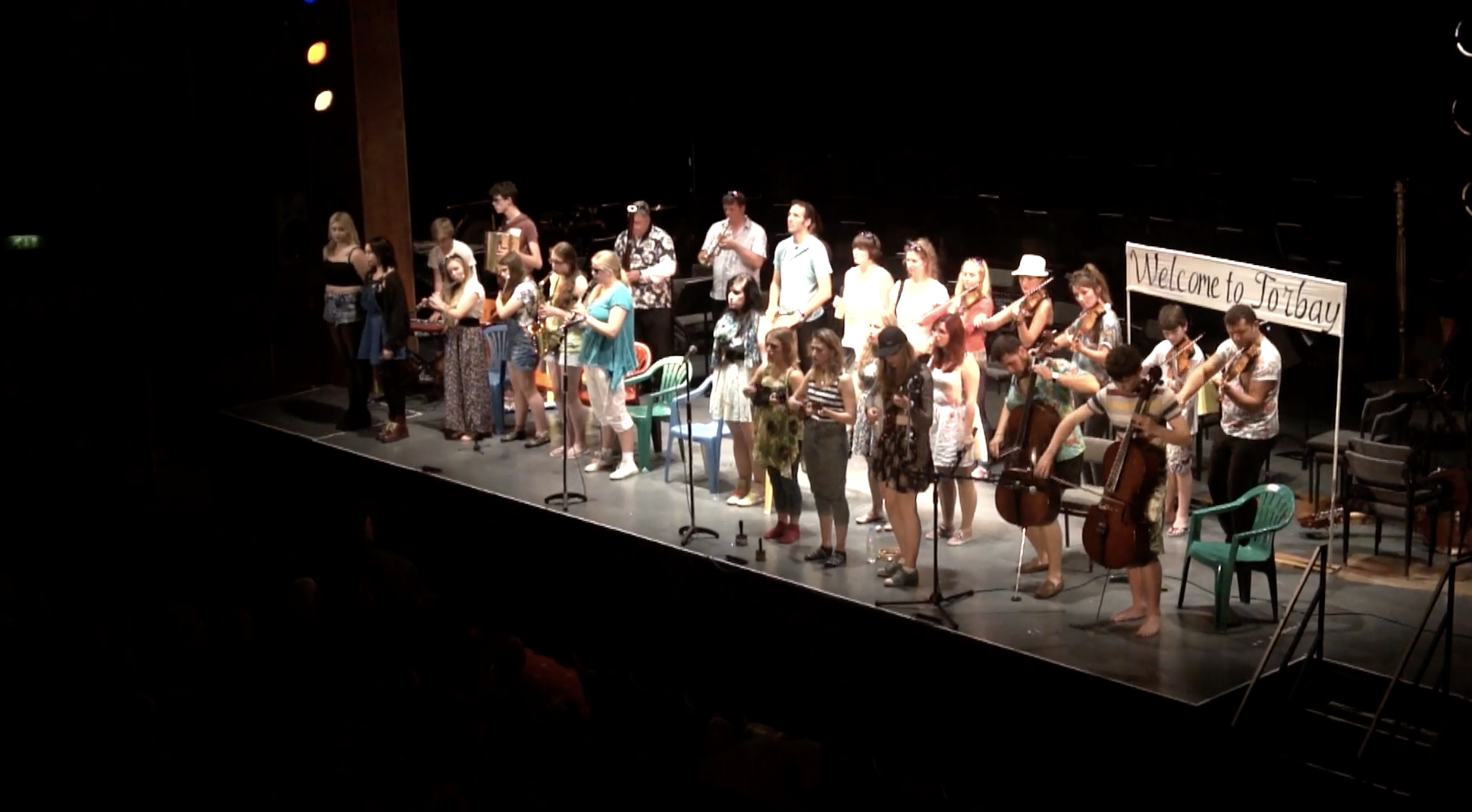 Group of musicians and singers performing on stage with a 'Welcome to Torbay' sign in the background.