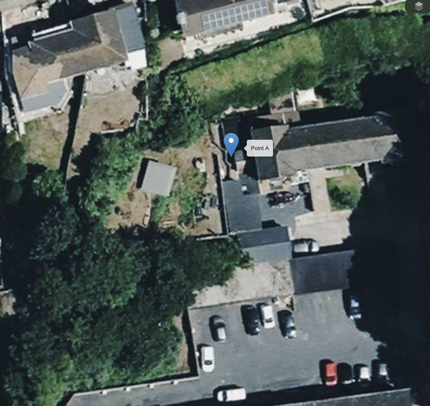 Aerial view of a small urban backyard with trees, a lawn, a paved path, a shed, and surrounding buildings, including parked cars along a parking lot.