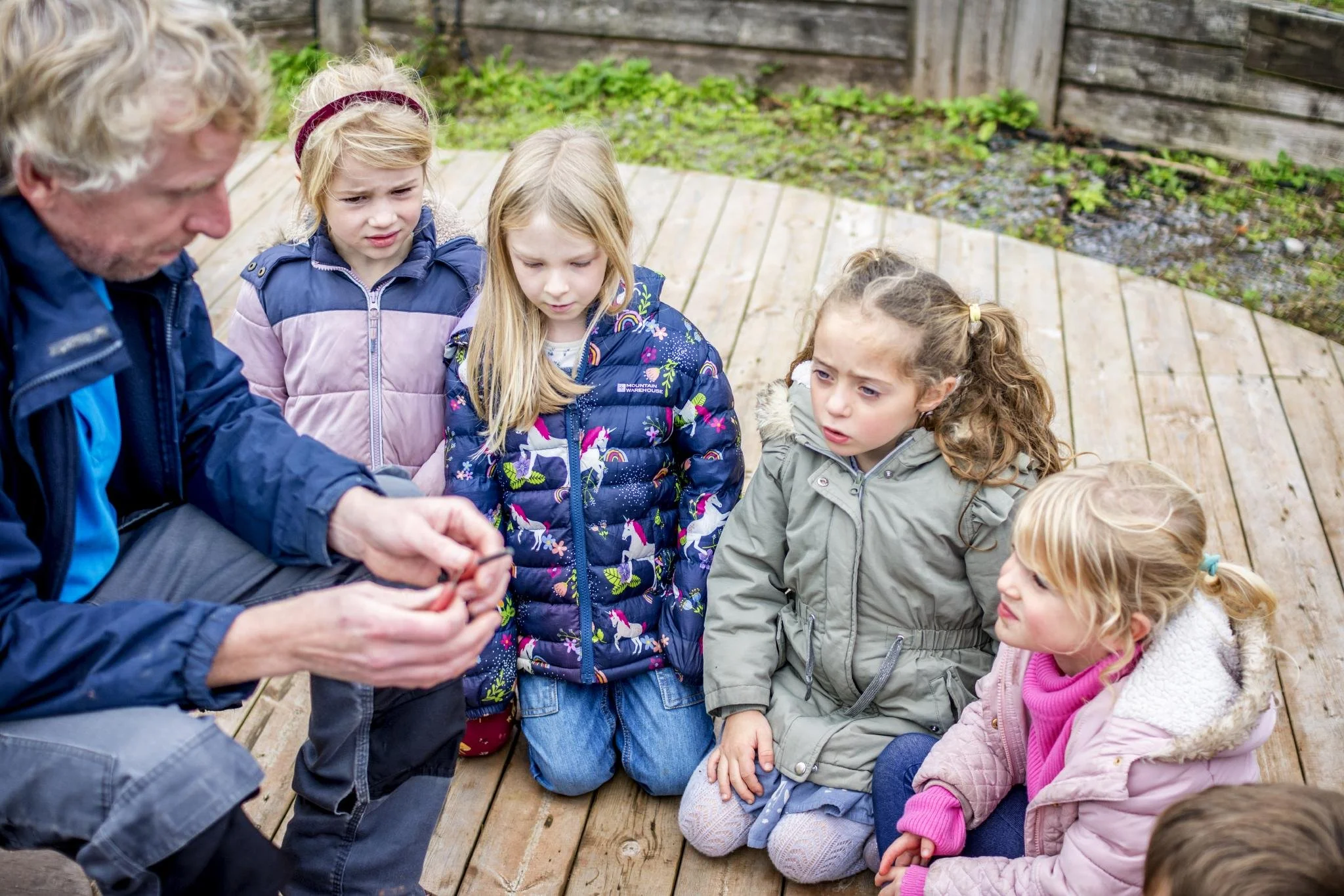 An older man with gray hair and a blue jacket shows something to a group of young girls sitting on a wooden deck outdoors, with a wooden fence and green plants in the background. The girls are wearing jackets and have focused expressions.