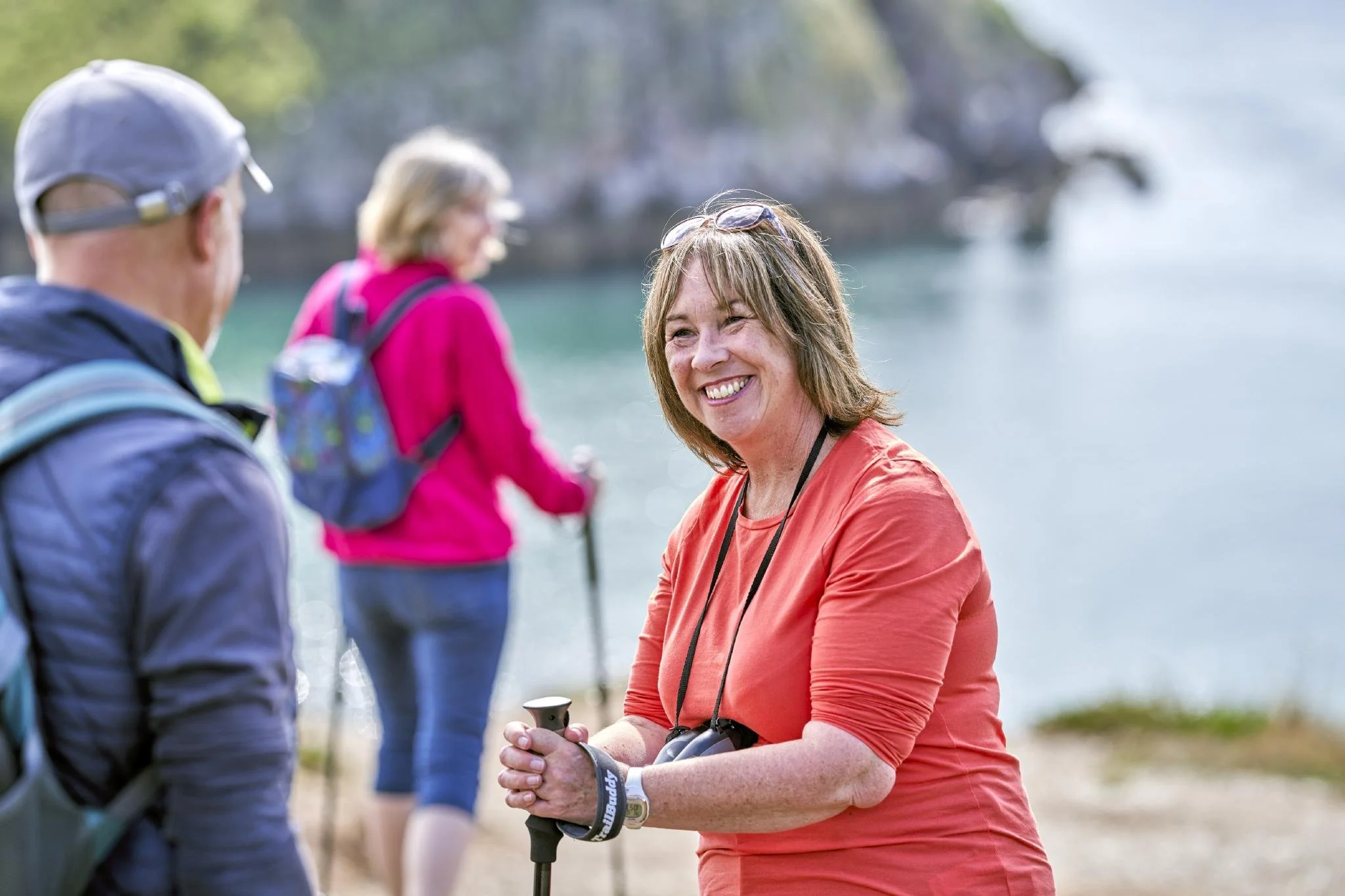 A woman smiling and talking to a man outdoors near a body of water, with other people in the background, dressed casually for a sunny day.