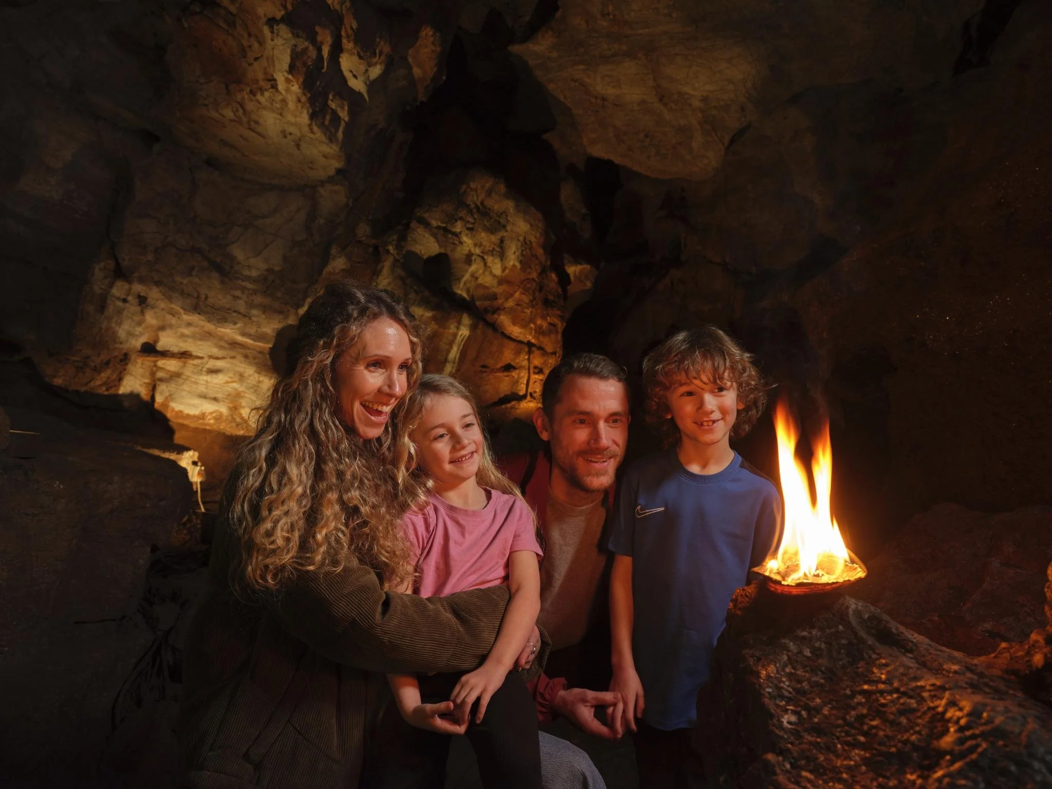 A group of five people, three children and two adults, smiling and sitting close to a small fire inside a cave with rocky walls and ceiling.