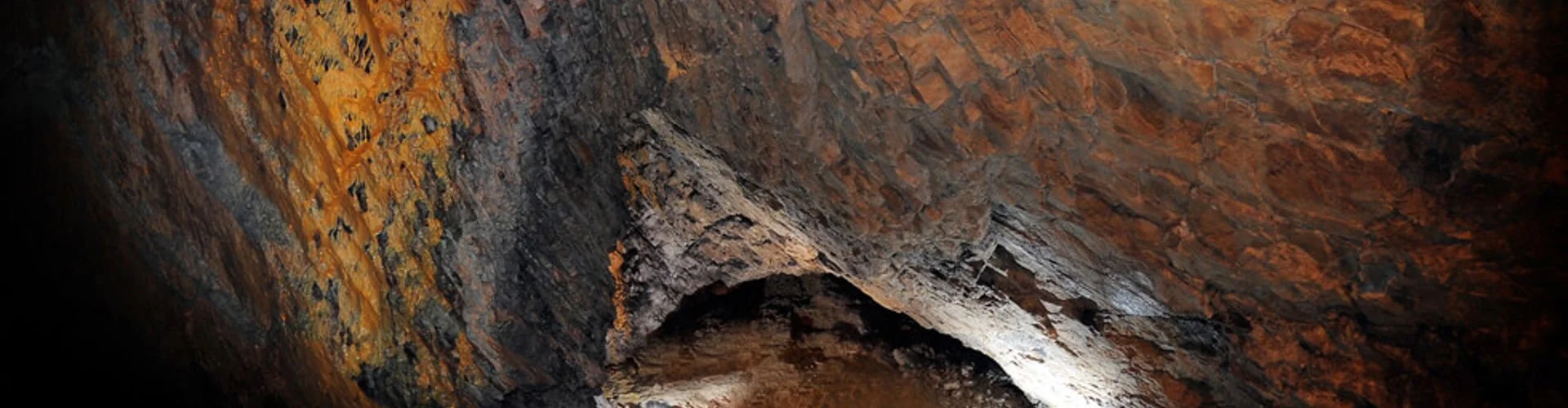 A view inside a dark, rocky cave with reddish-brown walls and a small opening.