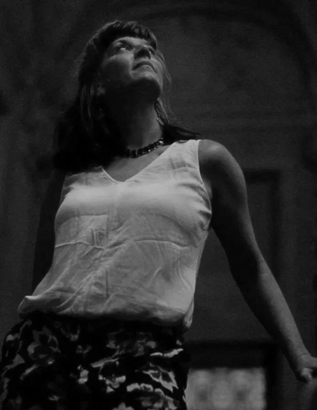 A woman with dark hair, wearing a sleeveless top and patterned skirt, looking upward with a serious expression.