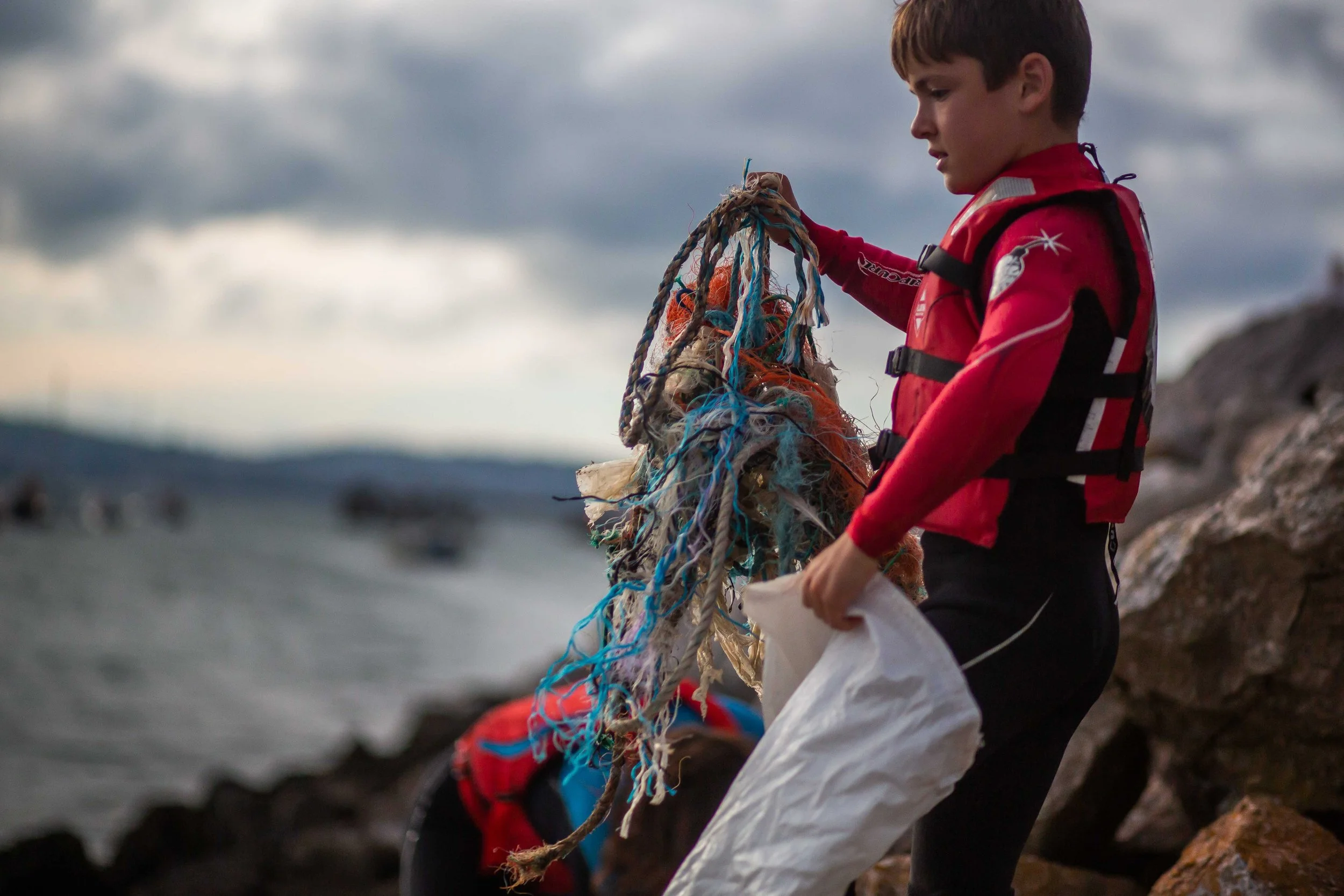 A young boy wearing a red waterproof jacket and black pants is outdoors on a rocky shoreline, holding a tangled fishing net with colorful ropes as clouds gather in the sky.