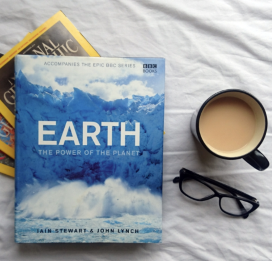 A book titled "Earth: The Power of the Planet" by Iain Stewart and John Lynch, a pair of glasses, a cup of coffee with cream, and some other magazines or books on a white surface.