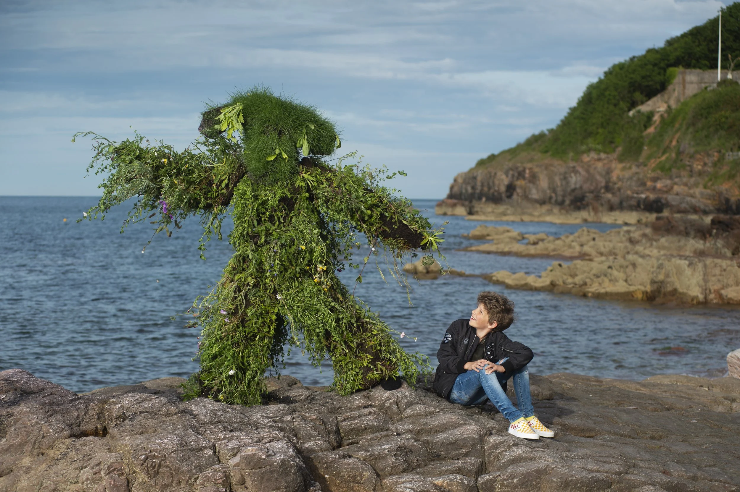 A young boy sitting on rocks by the water, smiling and looking at a large abstract sculpture made of greenery and flowers, with a rocky coastline and green hillside in the background.