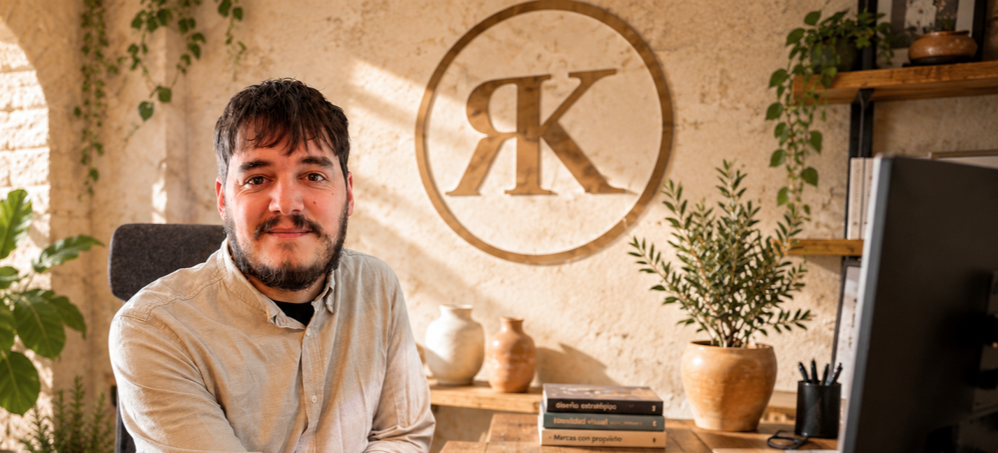 A man sits at a desk in an office with a large circular logo with two 'K's on the wall behind him, surrounded by potted plants and books.