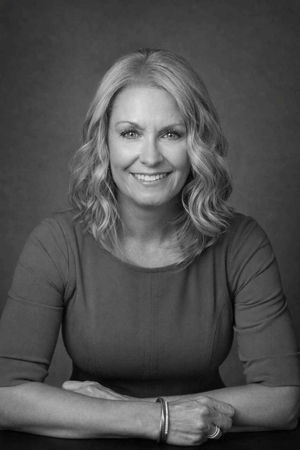 Black and white portrait of a woman with shoulder-length wavy hair, smiling, wearing a long-sleeved top, with a bracelet and ring on her left hand.