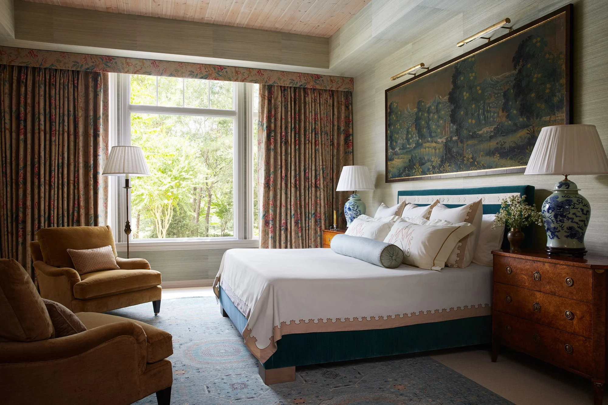 A cozy bedroom with a large window showing green trees outside. The room has floral curtains, a wooden bedside table with a blue and white vase lamp, a neatly made bed with white linens and decorative pillows, and a pair of armchairs near the window.