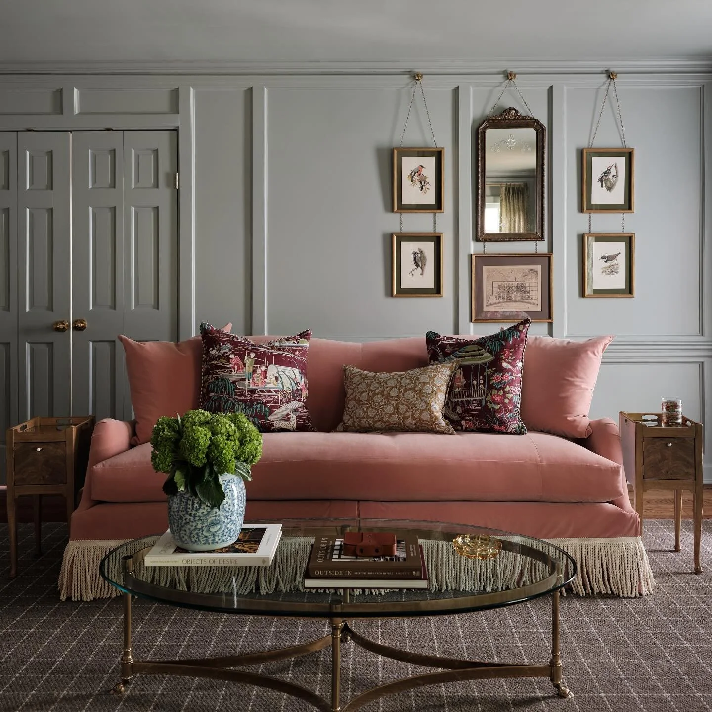 A hotel room with a red velvet sofa, multiple white pillows with colorful dot patterns, a blue upholstered headboard, a patterned rug, and a wall with multicolored circular accents. Two lamps are mounted above the headboard, and a set of curtains covers a window on the right side.