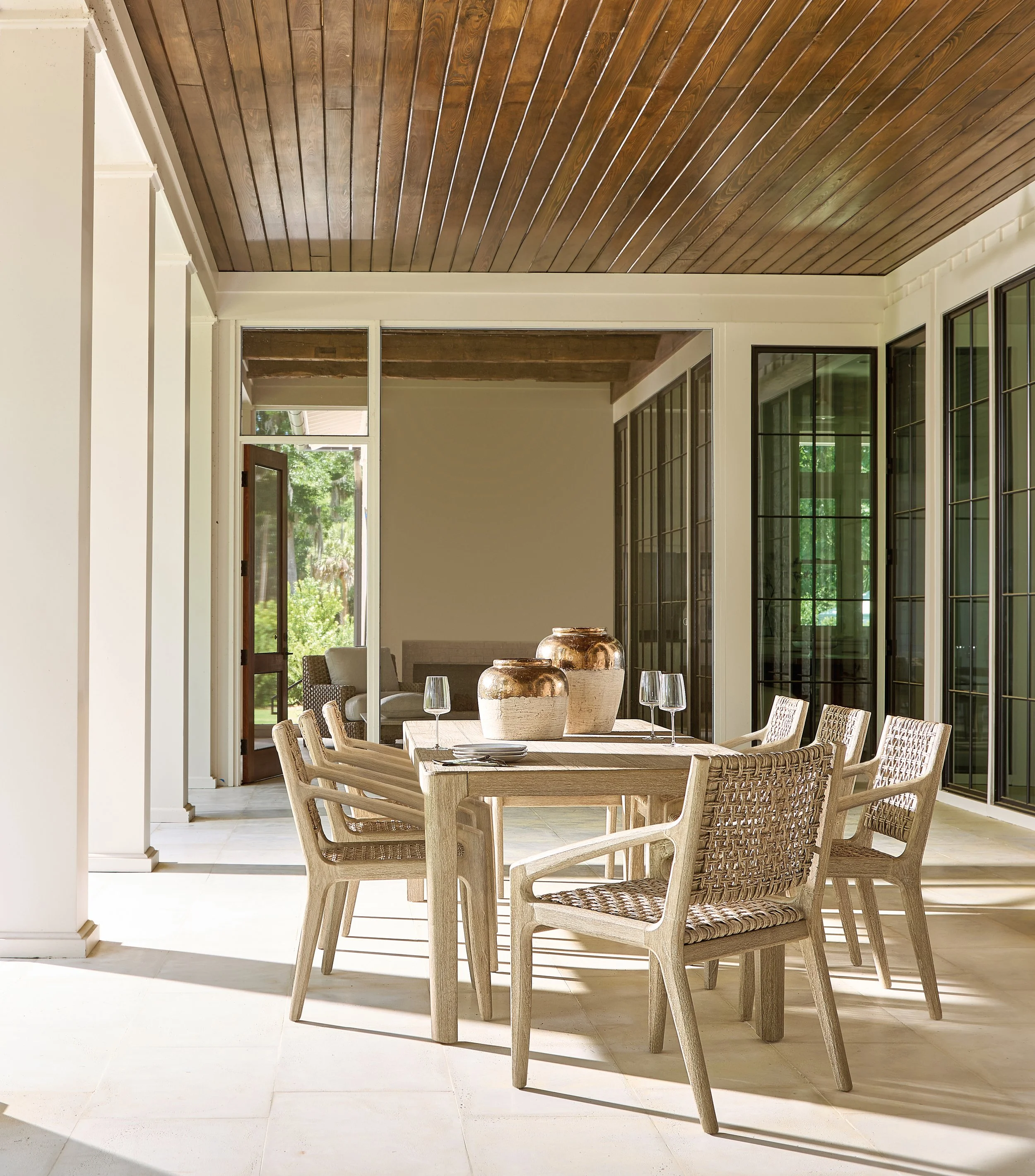 Dining area with wooden table, woven chairs, decorative pottery, glassware, and large windows with a view of greenery outside, sunlight casting shadows on light-colored tile flooring, wooden ceiling.