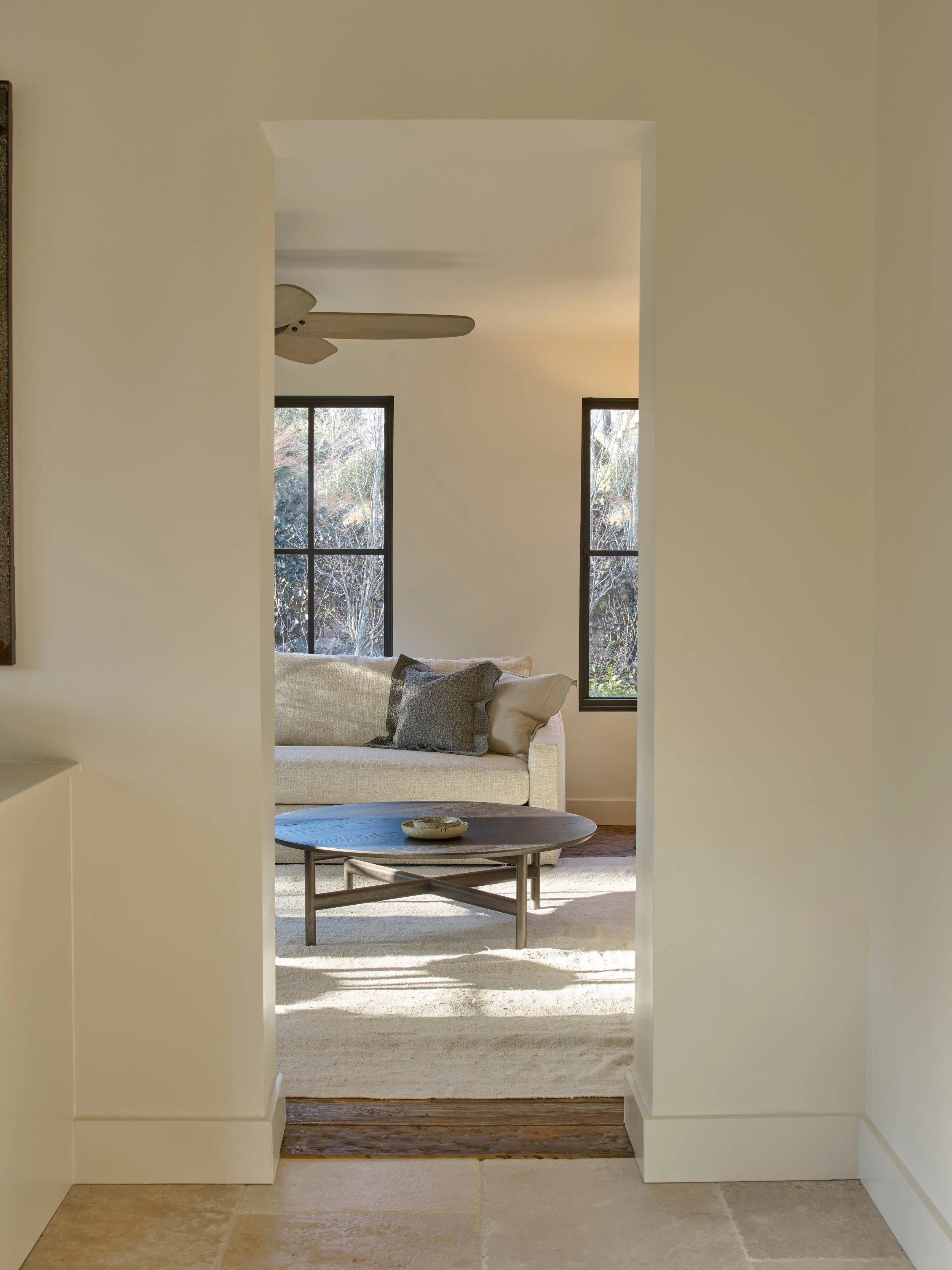 View through an archway into a living room with black framed windows showing trees outside, light-colored walls, a beige sofa with pillows, a round coffee table, and a ceiling fan.
