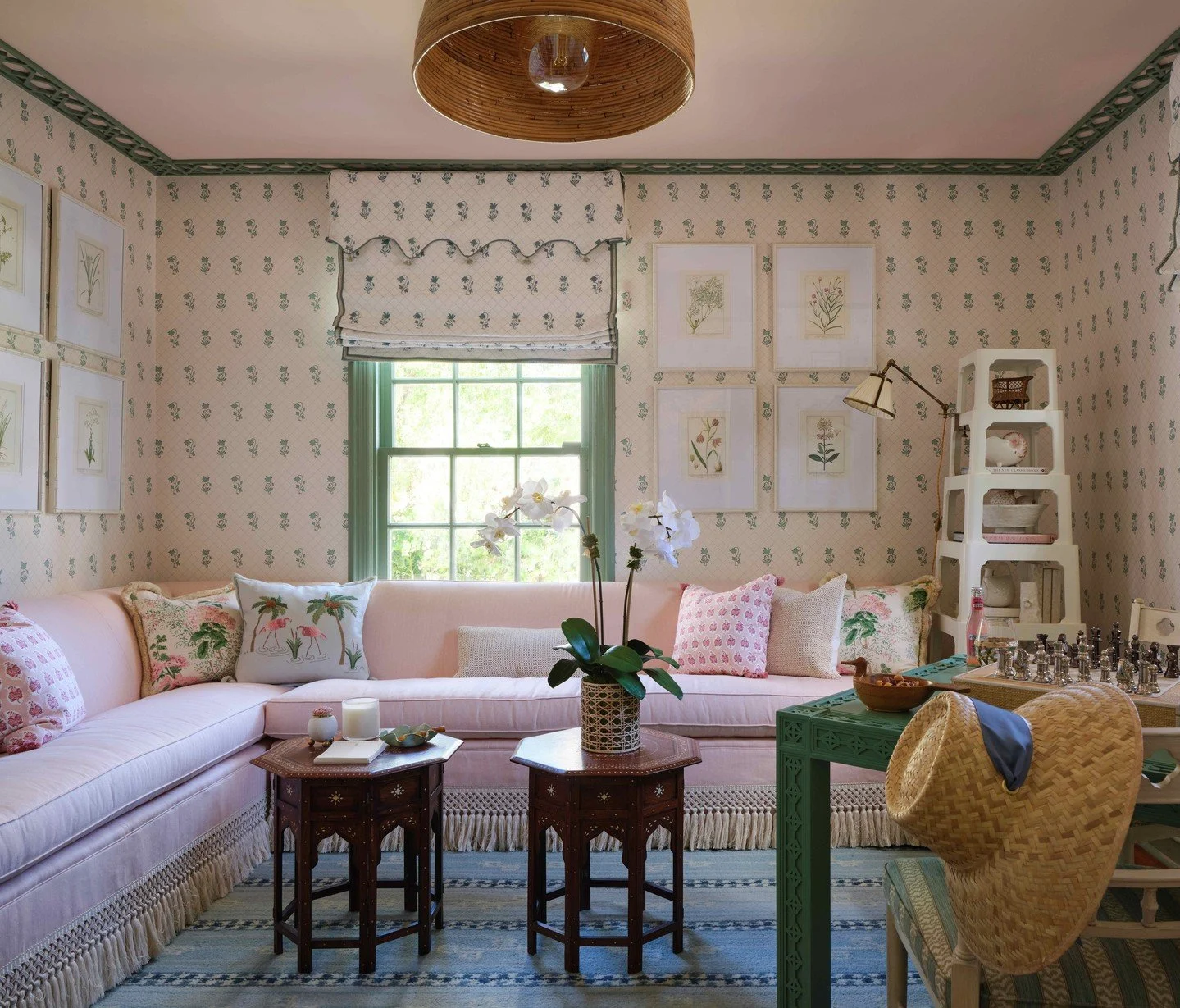 A room with blue and white checkered wallpaper and ceiling. The room contains various blue furniture, including a blue tufted sofa, a blue armchair, and a striped ottoman. There is a round wooden side table with books and a black desk lamp, a floor lamp with multiple black lampshades, and framed artwork on the walls. The window has checkered curtains matching the wallpaper, and there is a striped rug on the floor.