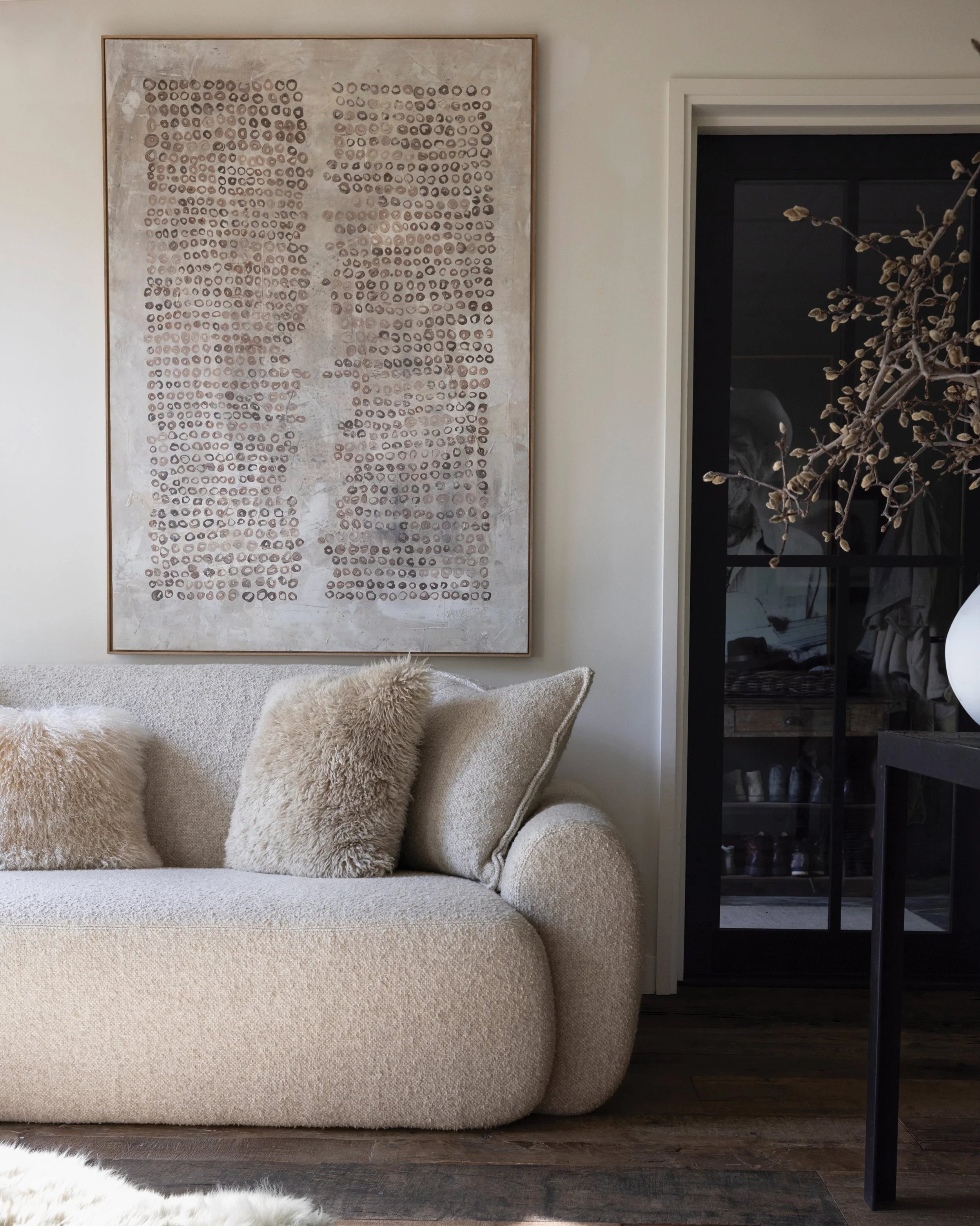 Living room with beige sofa and plush pillows, abstract wall art, and a black-framed glass cabinet with a dried branch arrangement.