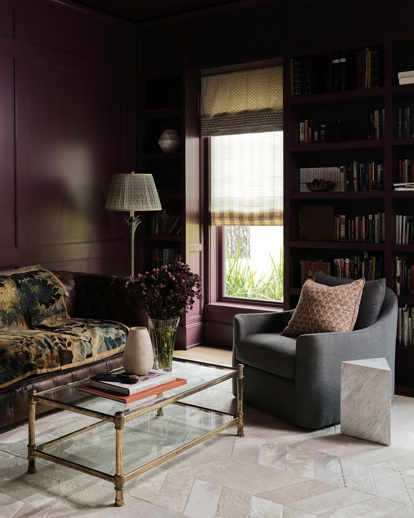 A room with blue and white checkered wallpaper and ceiling. The room contains various blue furniture, including a blue tufted sofa, a blue armchair, and a striped ottoman. There is a round wooden side table with books and a black desk lamp, a floor lamp with multiple black lampshades, and framed artwork on the walls. The window has checkered curtains matching the wallpaper, and there is a striped rug on the floor.