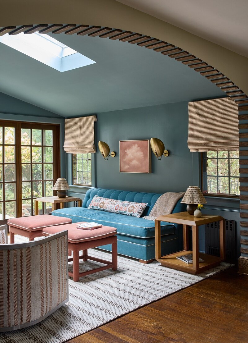 A cozy living room with a blue velvet sofa, pink ottomans, a wooden side table with a lamp, a patterned wall art, and windows with blinds. A skylight in the ceiling provides natural light, and the room features a mix of modern and vintage decor.