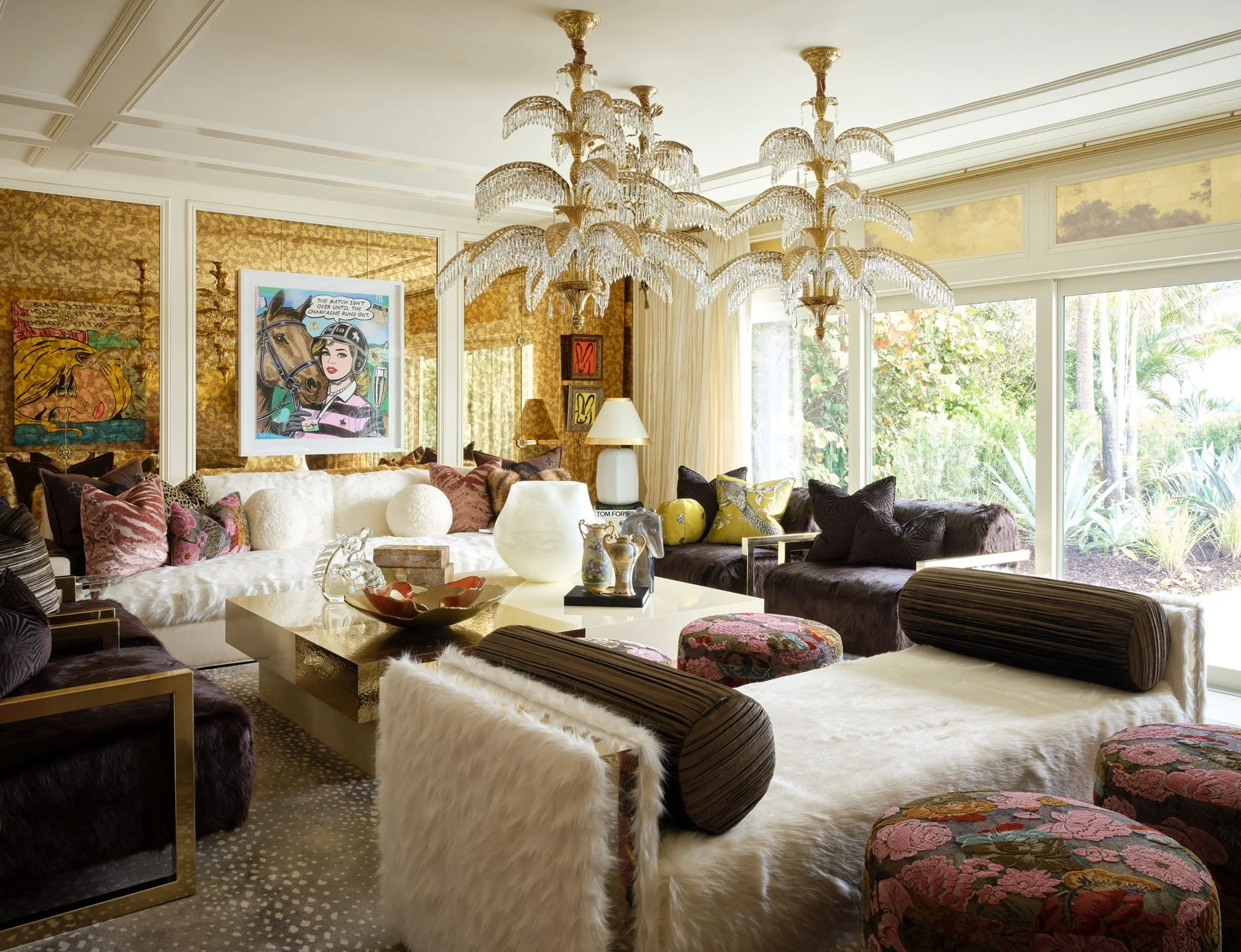 Luxurious living room with a large ornate crystal chandelier, colorful patterned pillows on plush sofas, and vibrant artwork on gold-accented walls with large windows revealing lush greenery outside.