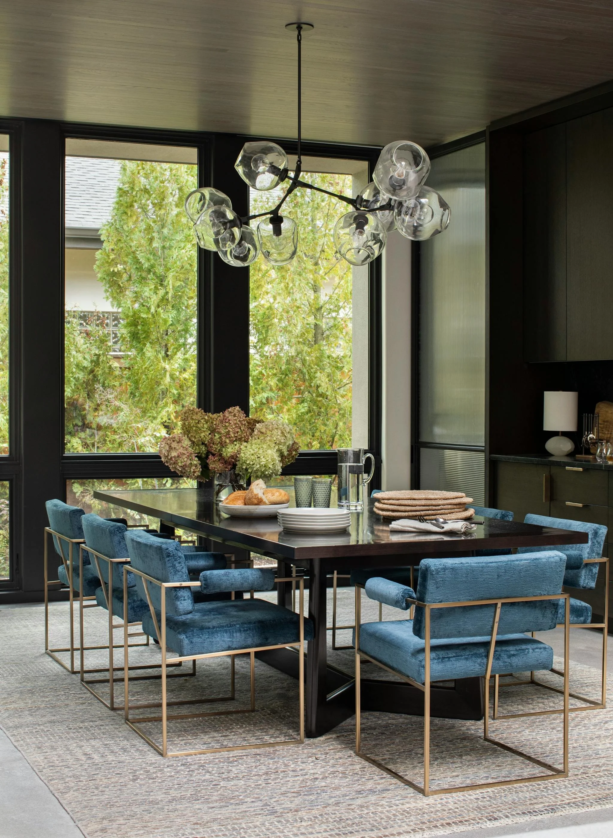 A modern dining room features a large black table surrounded by eight blue upholstered chairs with gold metal frames. On the table, there is a vase of hydrangeas, a loaf of bread on a plate, a glass pitcher of water, glasses, and stacked beige napkins. Large windows with black frames show green trees outside. A contemporary chandelier with multiple glass globes hangs above the table. The room has dark cabinetry and a neutral-colored area rug.