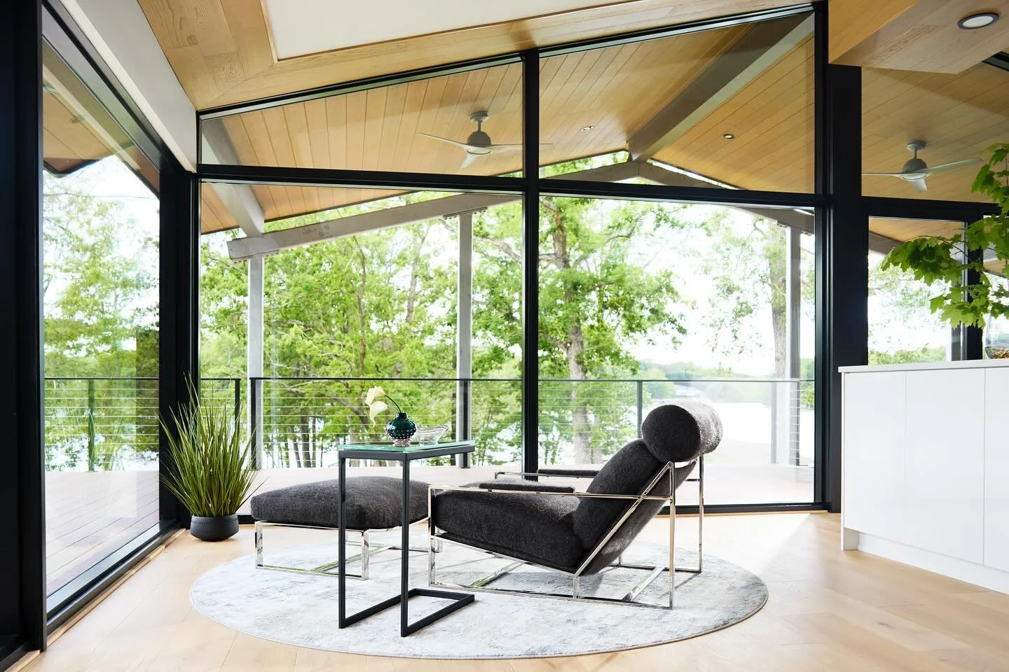 Modern living room with large glass windows, black-framed doors, a gray lounge chair, a side table with a small plant, an area rug, and a view of trees outside.