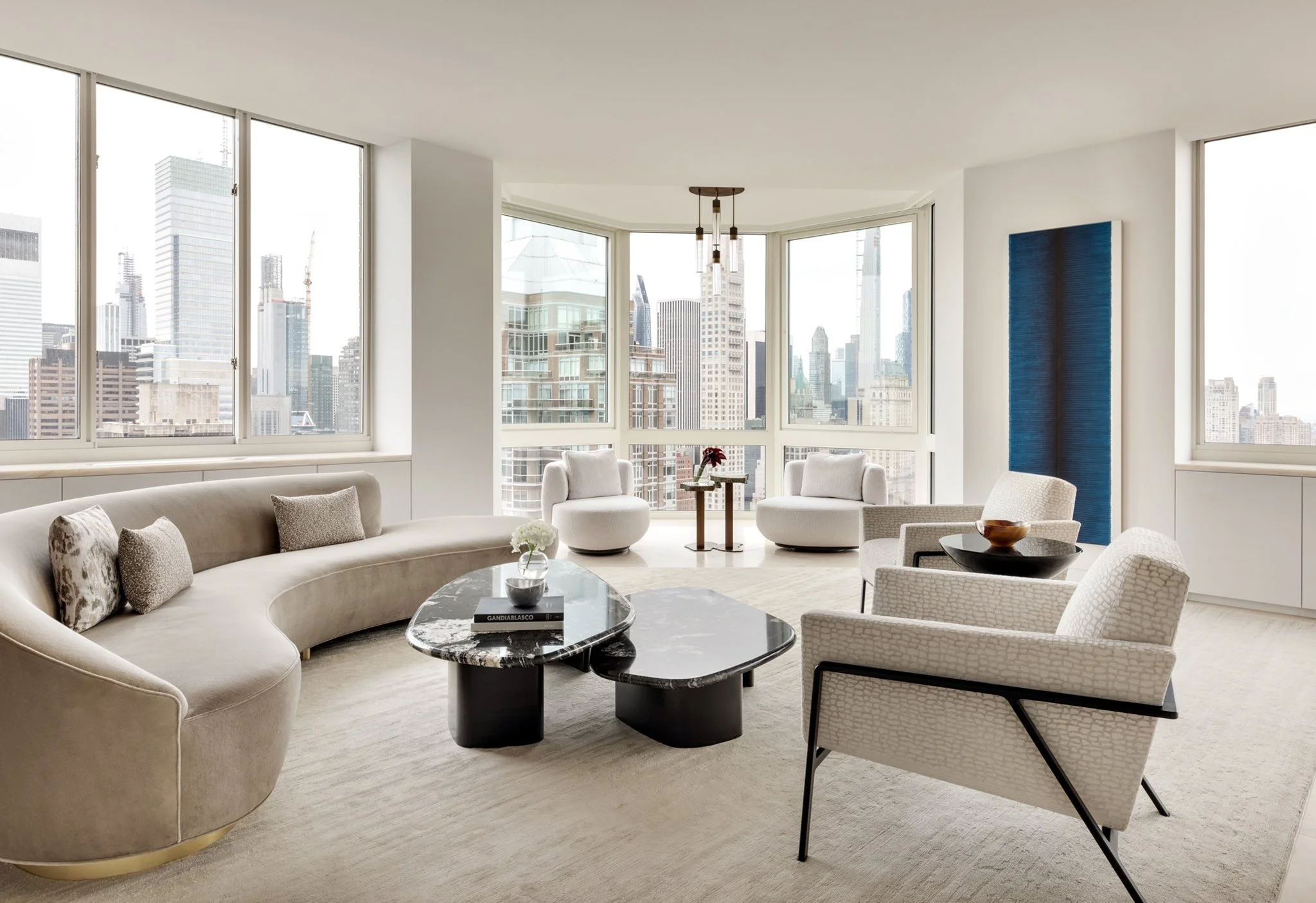Bright living room with large windows overlooking a city skyline, featuring a curved beige sofa, armchairs, a marble coffee table, and abstract artwork on the wall.