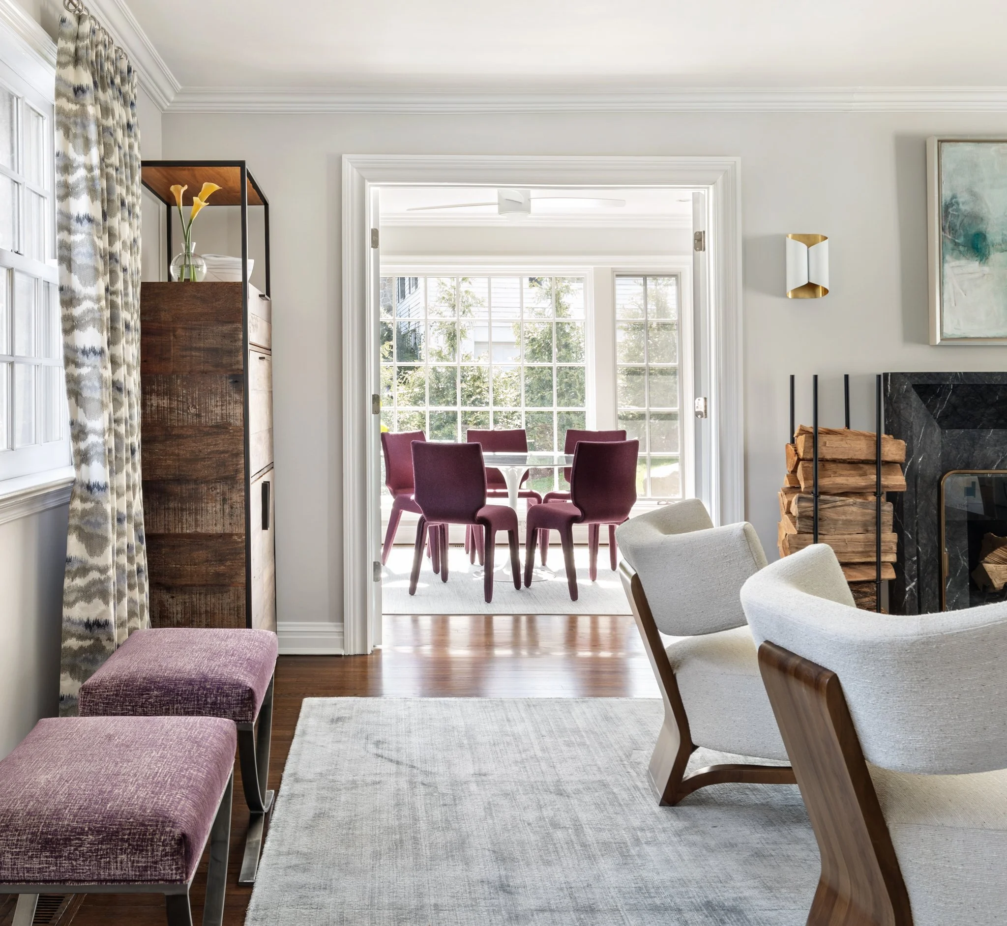 A view of a bright dining room with large windows, purple dining chairs around a table, a wooden bookshelf with a glass vase of yellow calla lilies, a fireplace with a black marble surround, a white armchair, a light grey rug, and a decorative wall sconce.