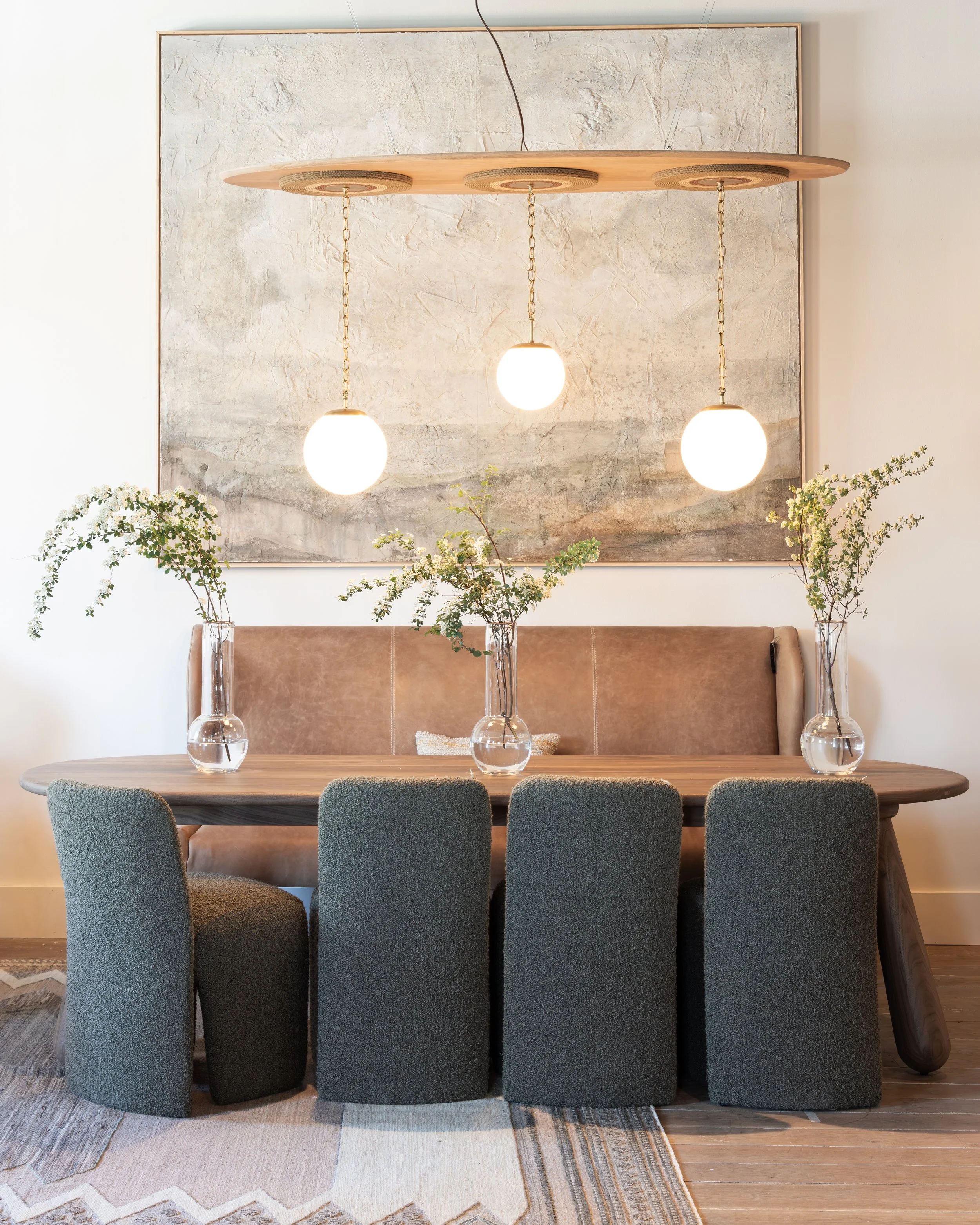 A dining room with a wooden table surrounded by six dark upholstered chairs. On the table, there are three glass vases with green leafy branches. Behind the table, a brown upholstered bench is against the wall. A large textured cream and gray abstract painting is hung on the wall above the bench. A modern wooden pendant light with three glowing globe bulbs hangs from the ceiling.
