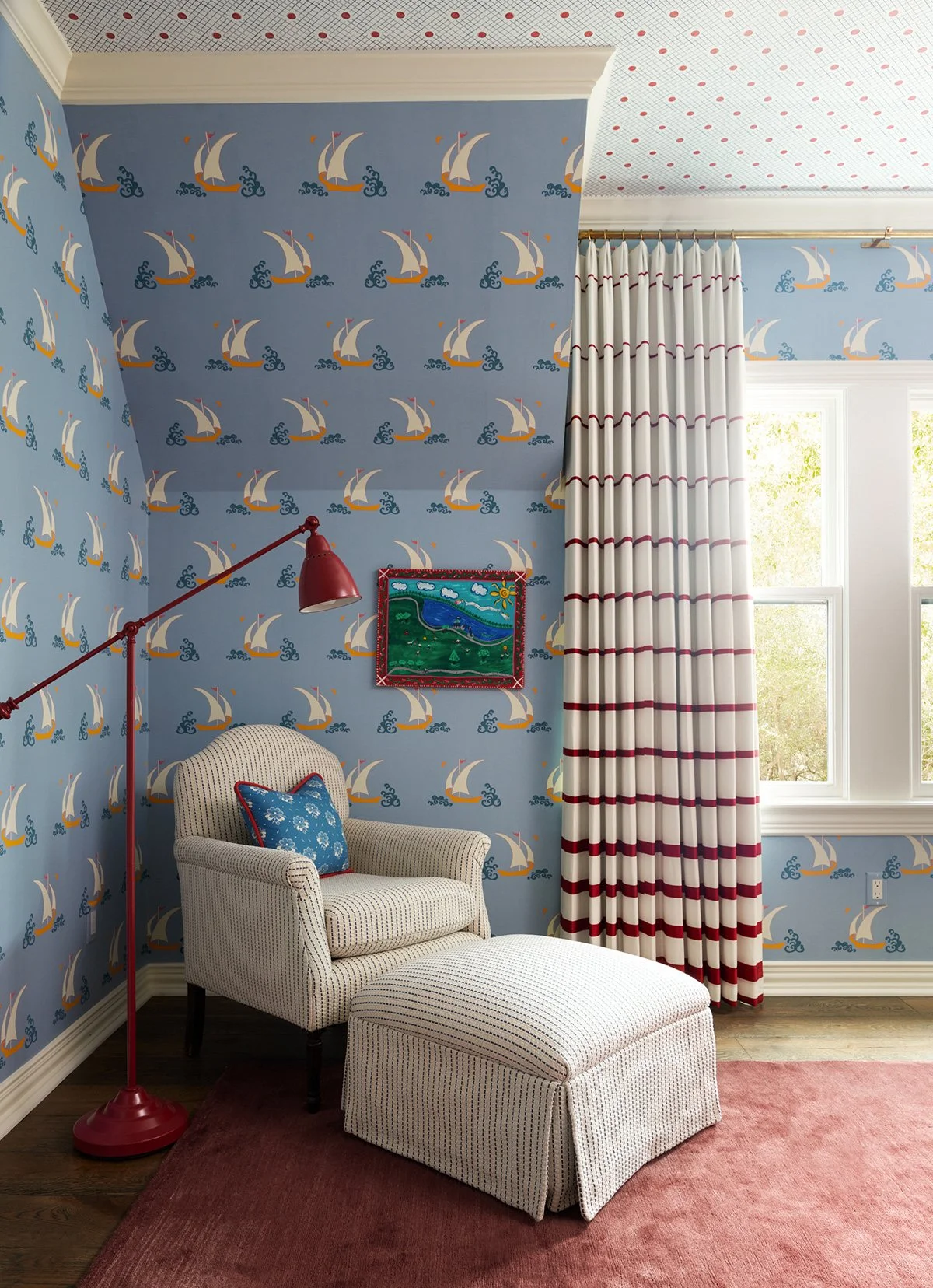 A cozy room with blue sailboat-themed wallpaper, a patterned white armchair with a matching ottoman, a red floor lamp, a small framed landscape painting, and striped red and white curtains. Light from a window illuminates the space.