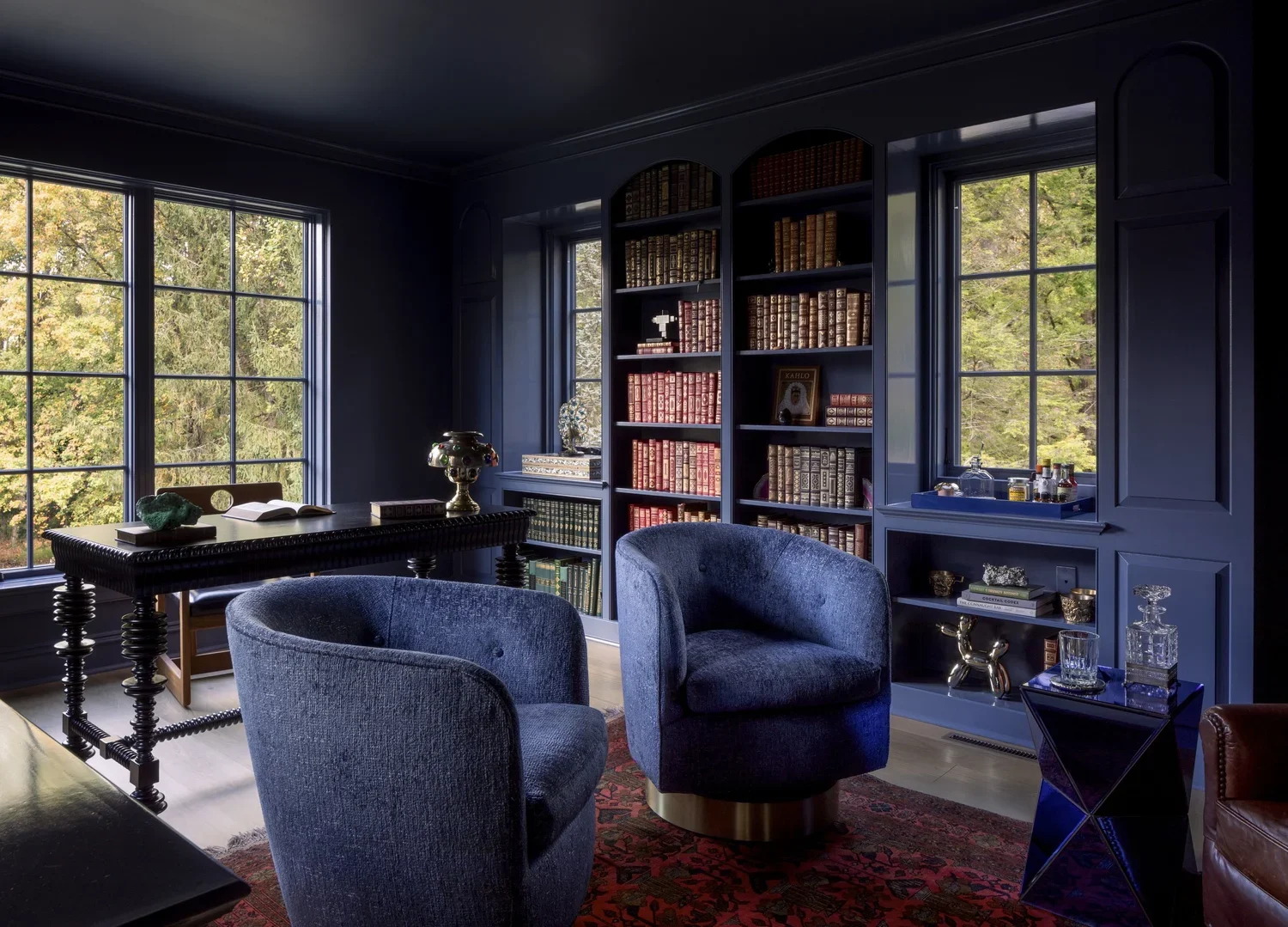 A cozy study room with dark blue walls and large windows showing green trees outside. The room features blue velvet armchairs, a dark wood table with decorative items, and a large bookshelf filled with leather-bound books. There are various decorative objects and glassware on a side shelf and table.