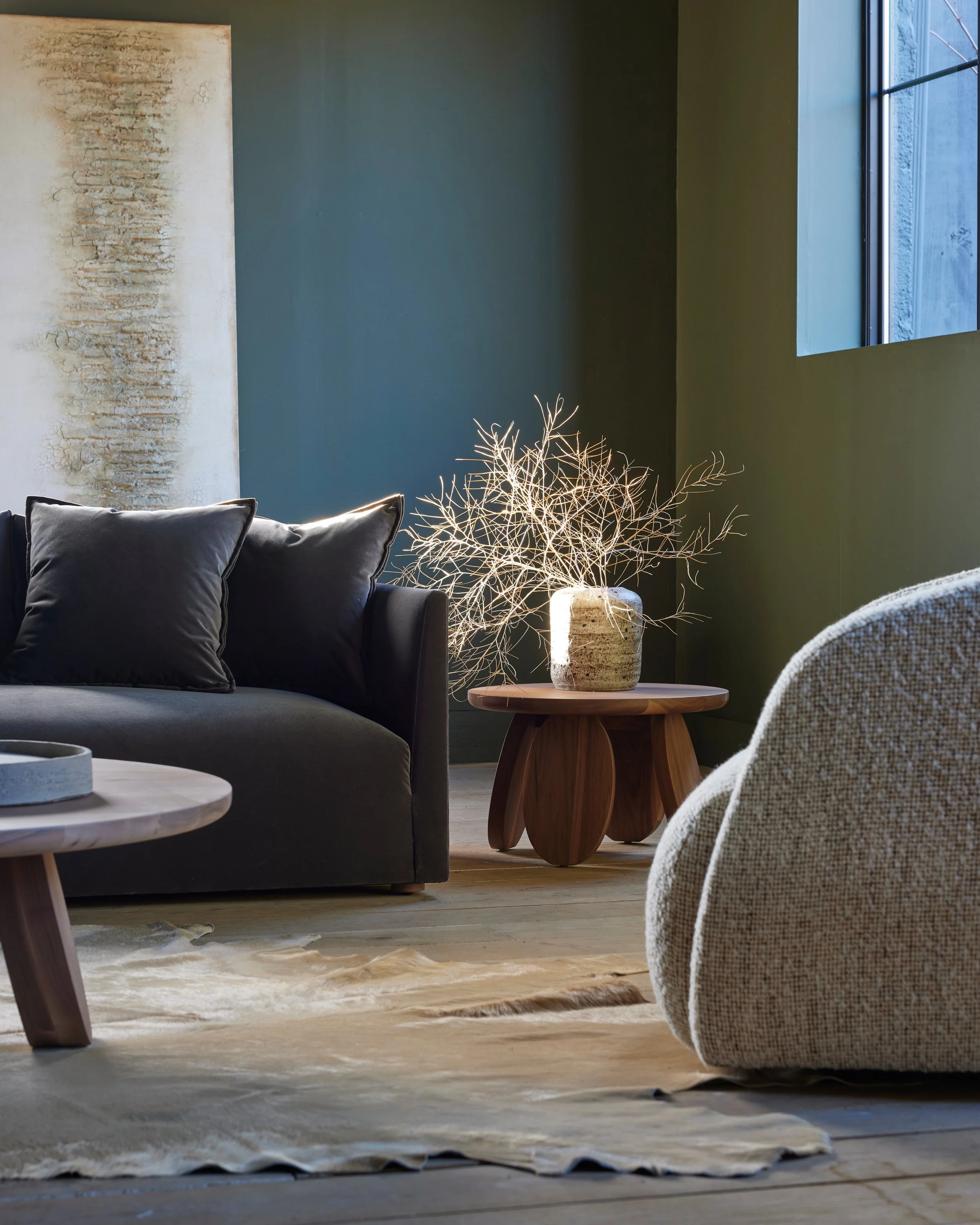 A living room with a dark gray sofa with pillows, a wooden side table with a decorative lamp, a textured beige area rug, a modern vase with dried branches, a large abstract wall art, a window allowing natural light, and a textured light-colored blanket on a chair.