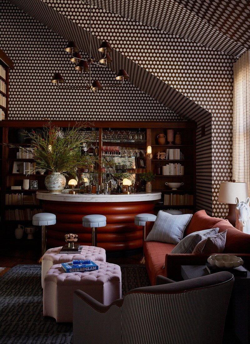 Interior of a cozy living space with a built-in bar, featuring patterned wallpaper, a ceiling light fixture, shelves with books and decorative items, a sofa with pillows, a tufted ottoman with snacks, a plush armchair, and a window with curtains.