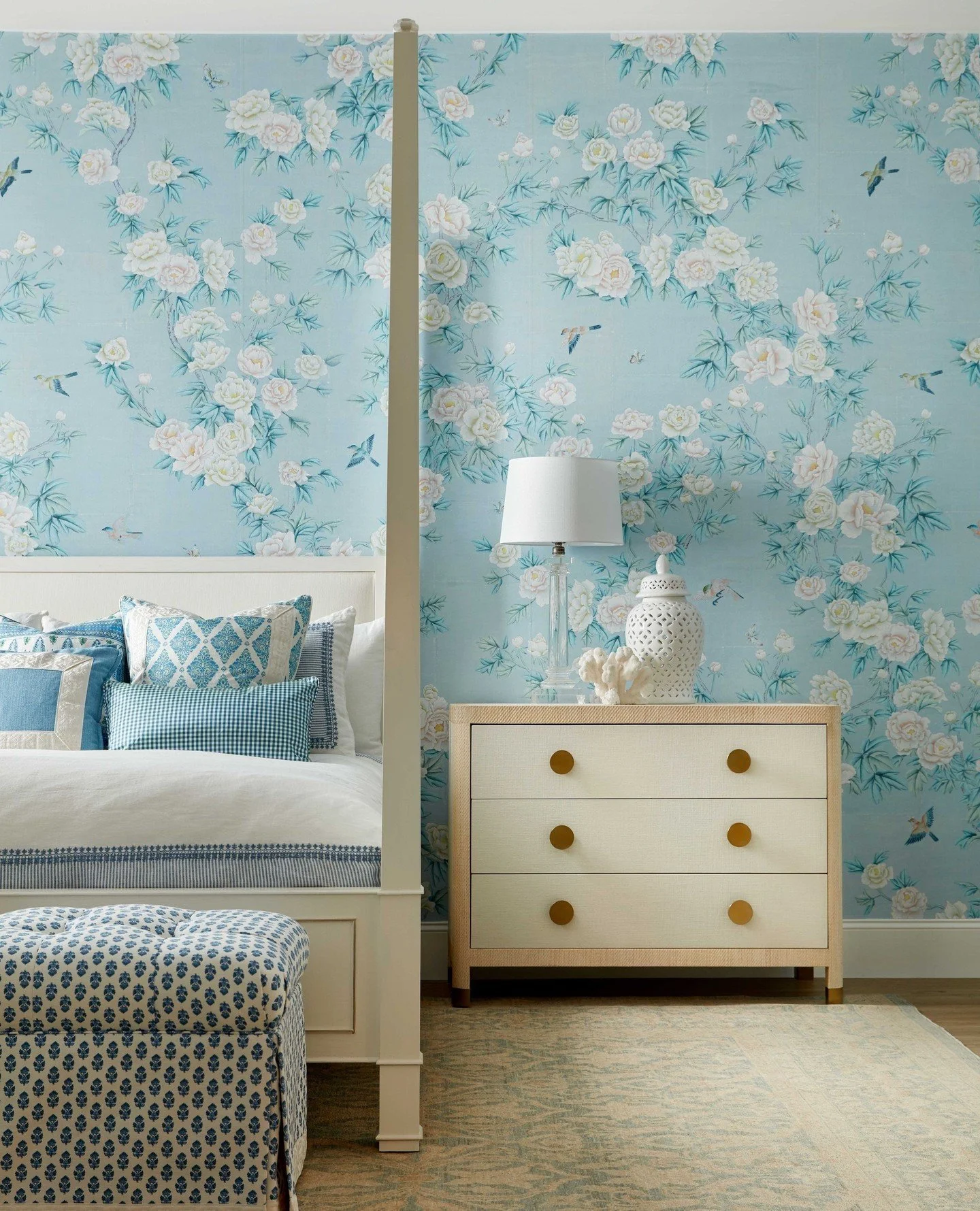 A hotel room with a red velvet sofa, multiple white pillows with colorful dot patterns, a blue upholstered headboard, a patterned rug, and a wall with multicolored circular accents. Two lamps are mounted above the headboard, and a set of curtains covers a window on the right side.