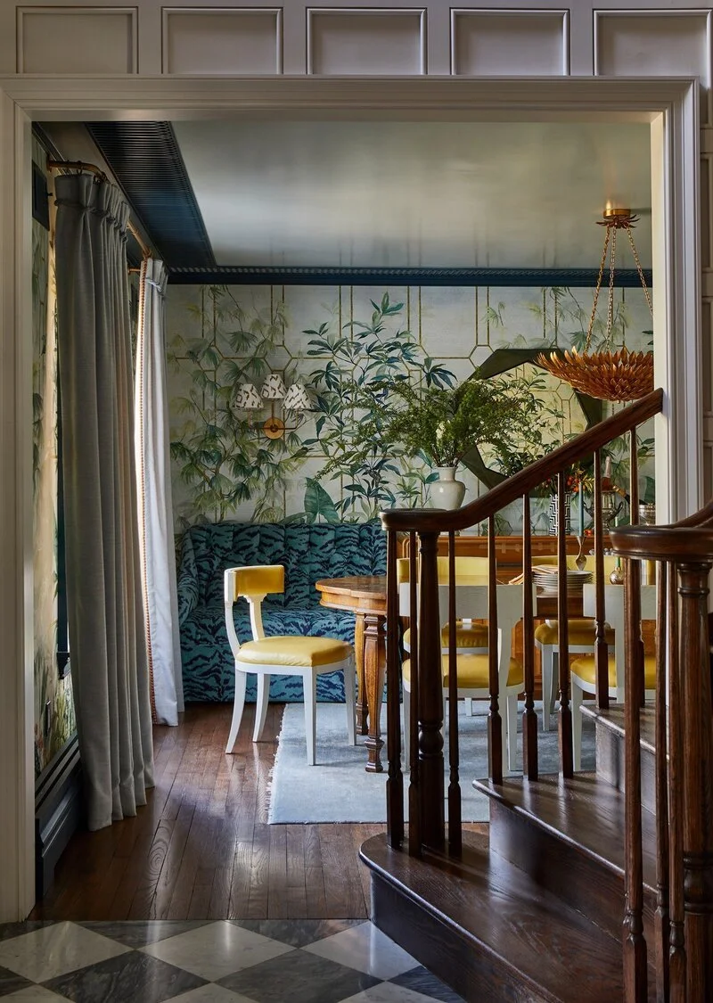 A cozy dining area with a blue patterned sofa, a yellow and white chair, a wooden dining table, and a large vase with green foliage. The wall features a wallpaper with a botanical design. There is a hexagonal mirror and a pendant light with a flower-shaped lampshade. Curtains cover the window, and a staircase with dark wood railing is visible in the foreground.