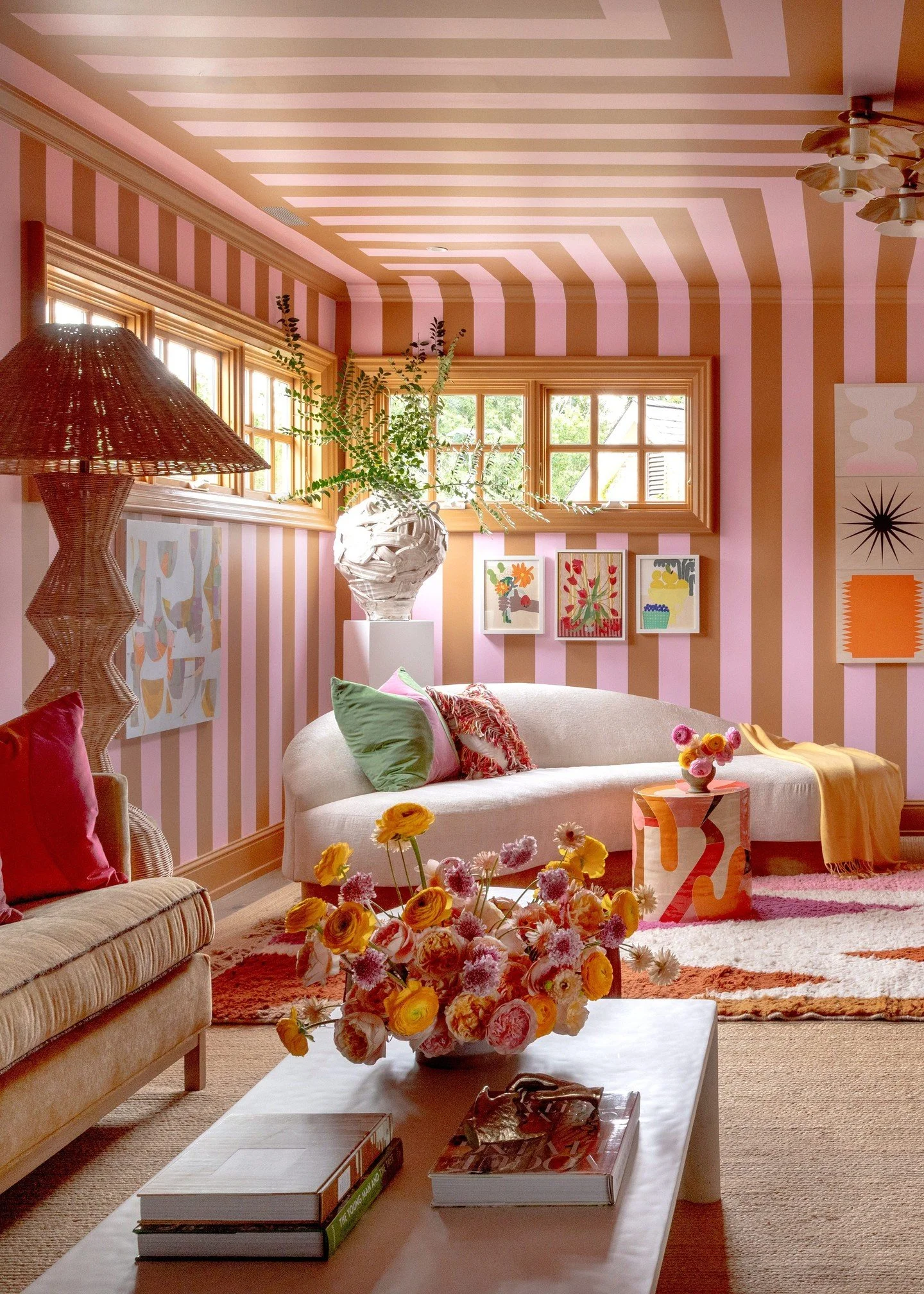 A room with blue and white checkered wallpaper and ceiling. The room contains various blue furniture, including a blue tufted sofa, a blue armchair, and a striped ottoman. There is a round wooden side table with books and a black desk lamp, a floor lamp with multiple black lampshades, and framed artwork on the walls. The window has checkered curtains matching the wallpaper, and there is a striped rug on the floor.