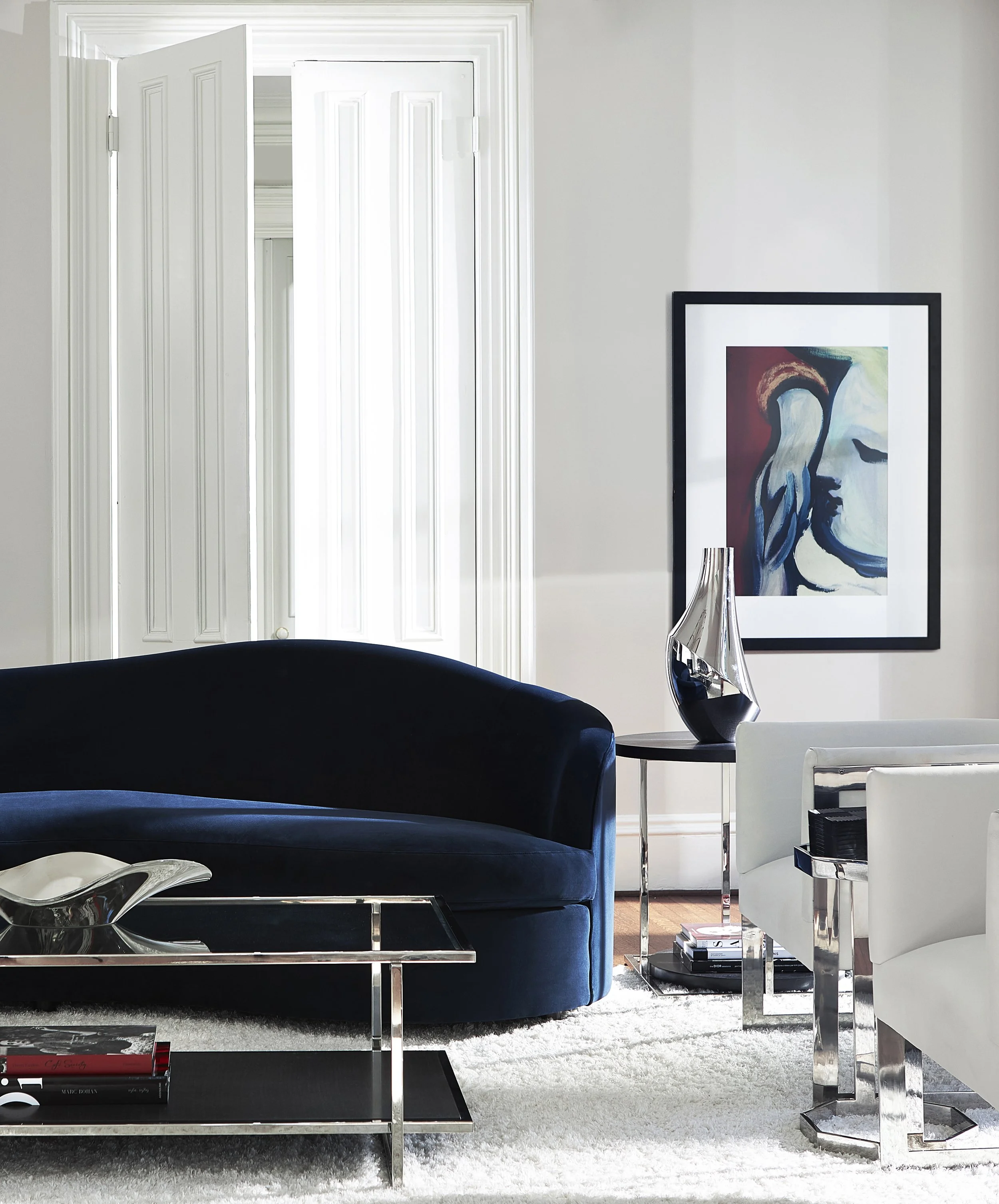 Modern living room with white walls, open window with white shutters, black velvet sofa, silver coffee table, abstract artwork in a black frame, decorative glass sculpture, white side table with magazines, and beige carpet.
