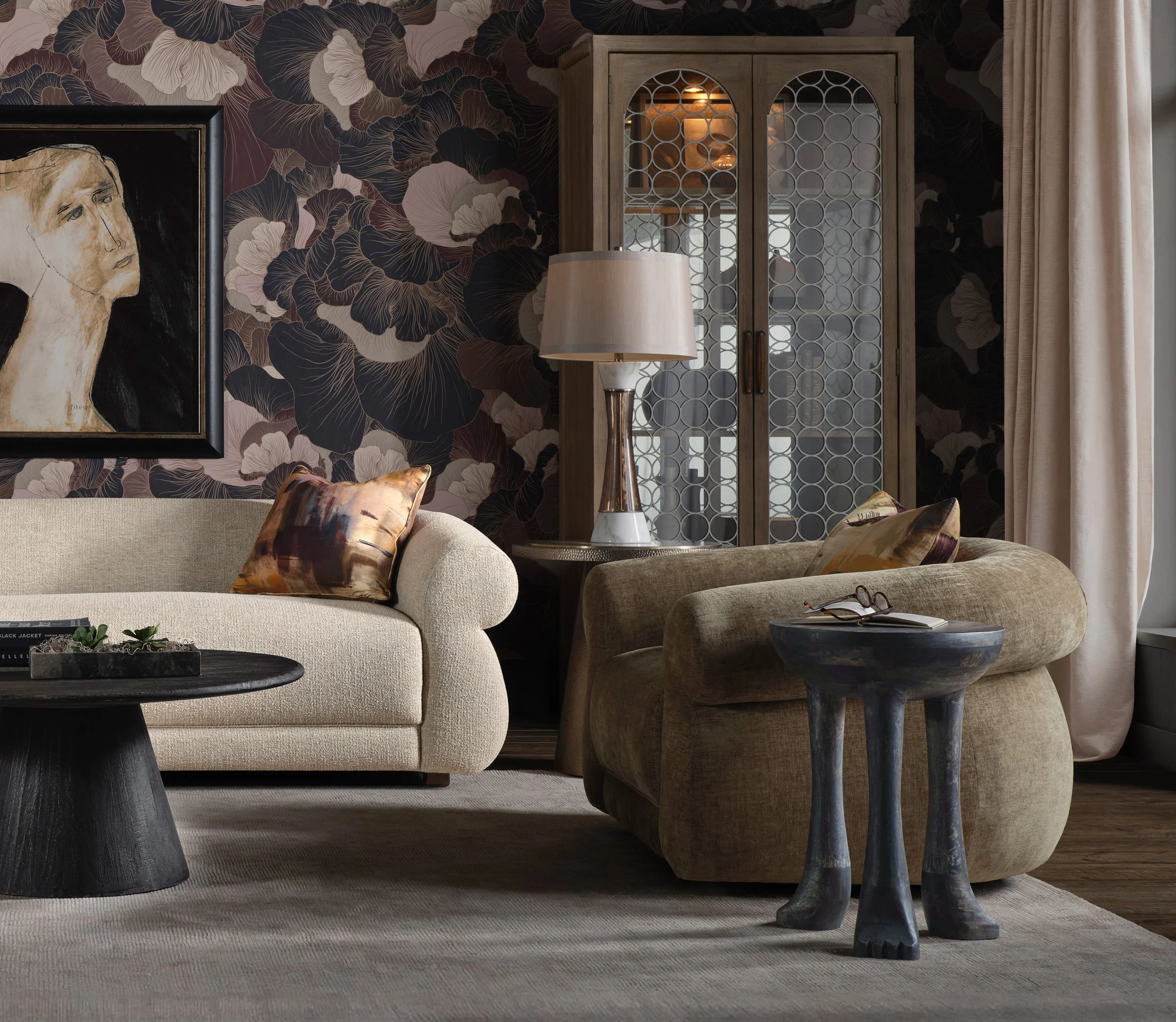 A living room with a white sofa and dark throw pillows, a round glass coffee table with magazines and decorative balls, and a fireplace. A standing lamp and a window with a view of trees under a clear sky are also visible.