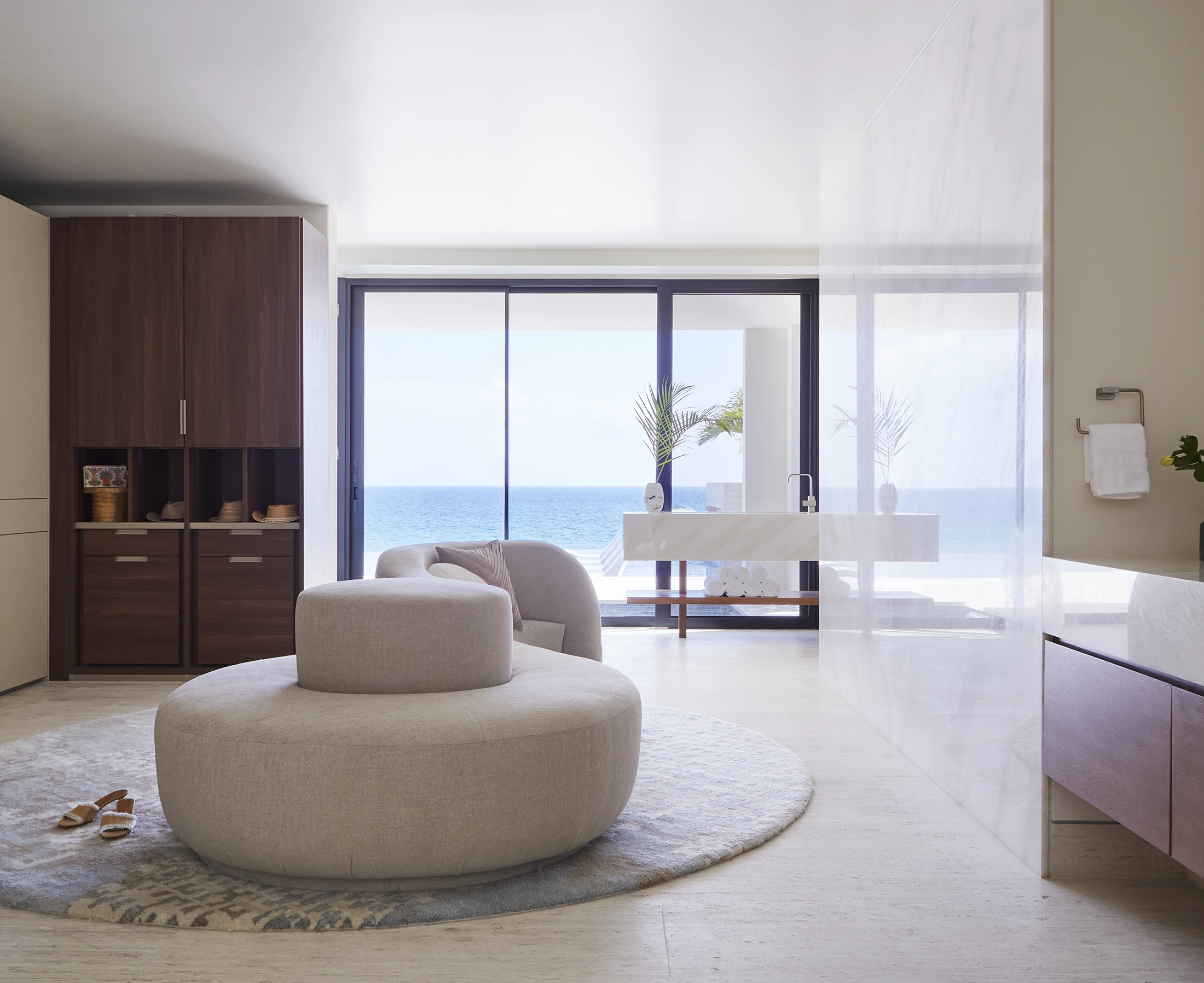 Living room with beige circular ottoman, plush armchair, and large window with ocean view, plants, and sheer curtains.