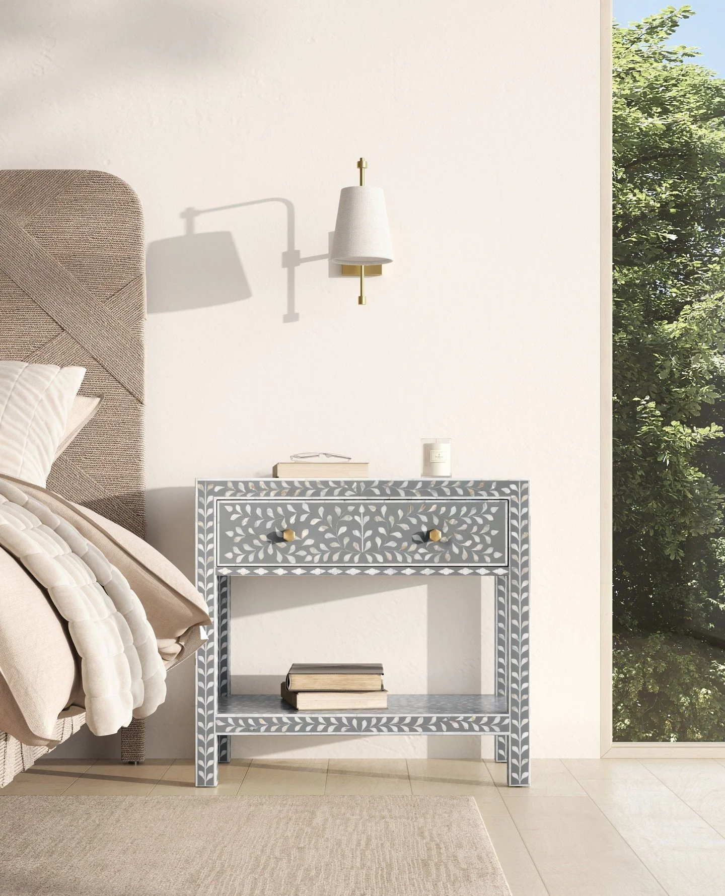 A bedroom corner with a beige bed and headboard, a gray decorative side table with books, a white candle, a white lamp with brass accents attached to a white wall, and a large window showing green trees outside.