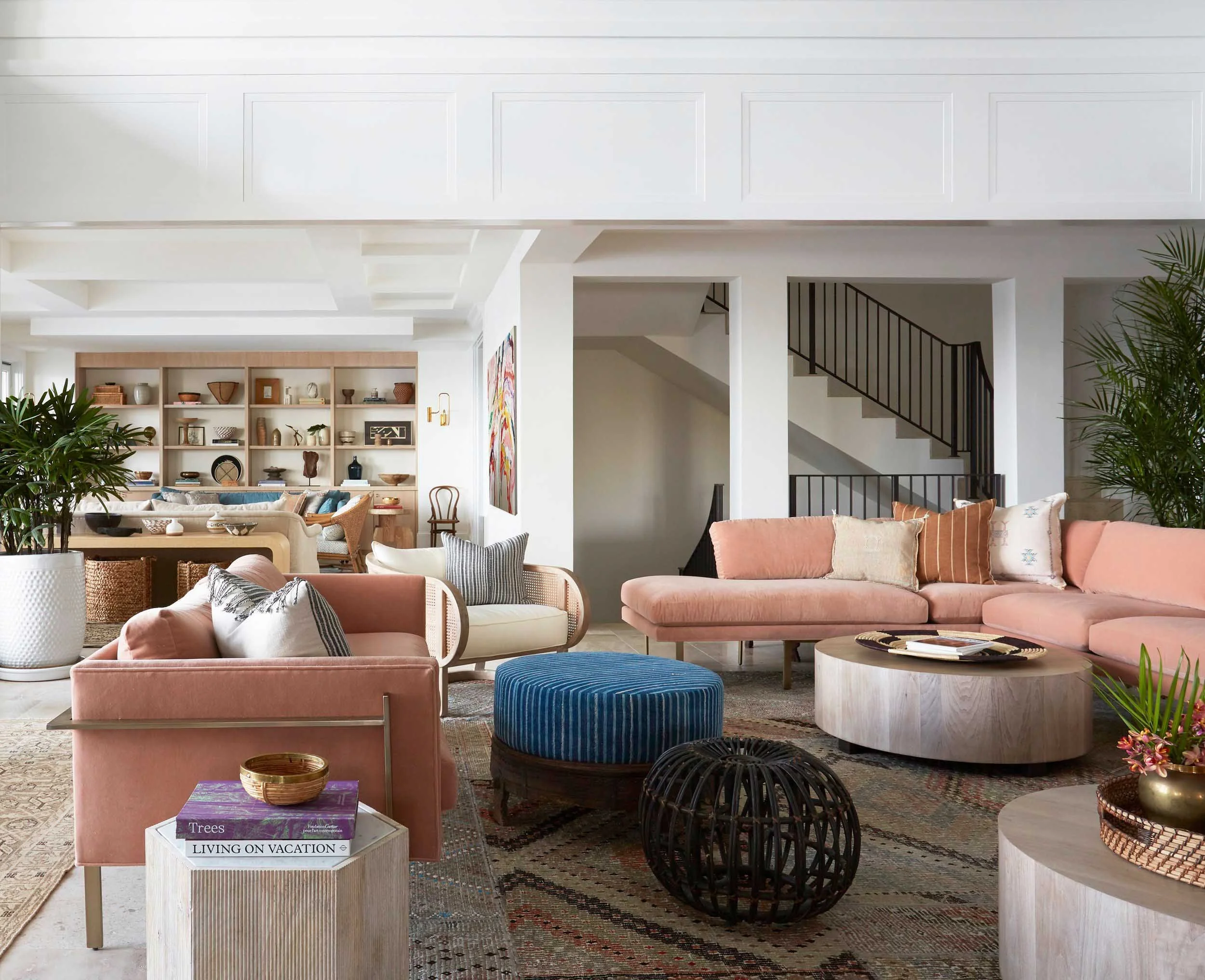 Bright living room with pink and beige furniture, wooden bookshelf filled with decorative items, plants, and colorful artwork.