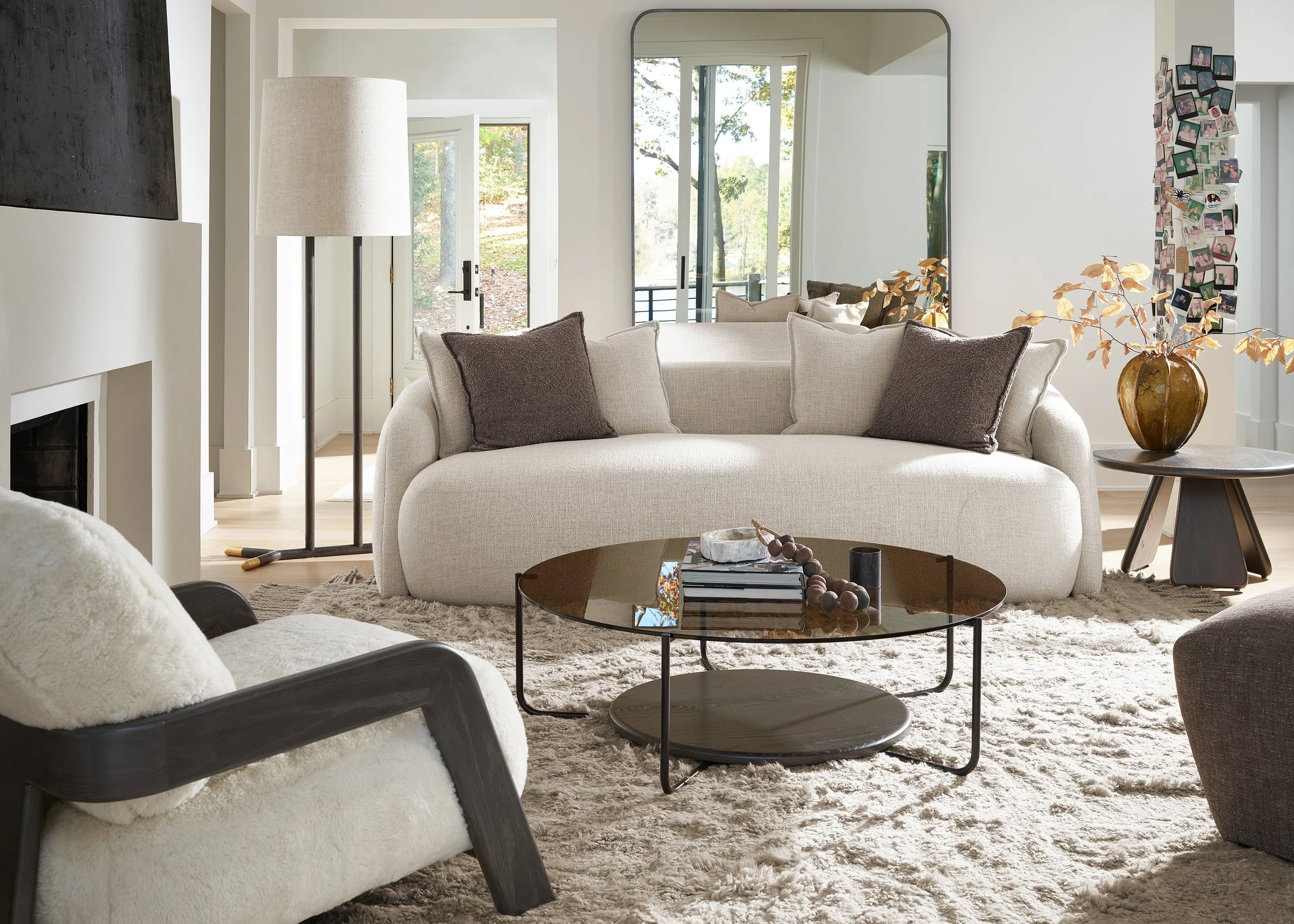 Interior of a modern living room with a white curved sofa, brown cushions, a black round coffee table with magazines and a beaded decoration, a black armchair with a white fur throw, a tall lamp, a large mirror, a collage of photos on the wall, and glass doors leading to an outdoor area with trees.