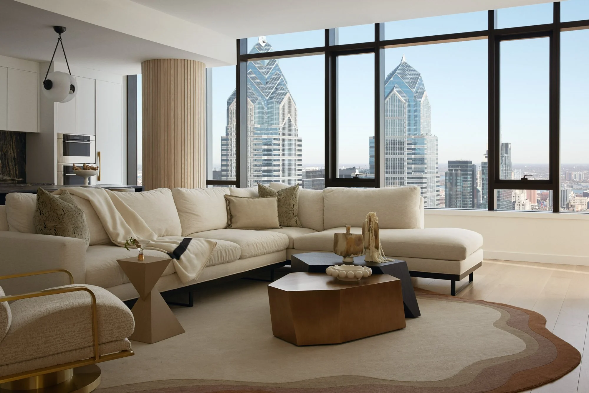 Modern living room with large floor-to-ceiling windows showcasing a city skyline with tall skyscrapers. The room has a cream-colored sectional sofa with decorative pillows, a wooden and black coffee table, and stylish side tables. There is a large area rug with a curve border pattern.